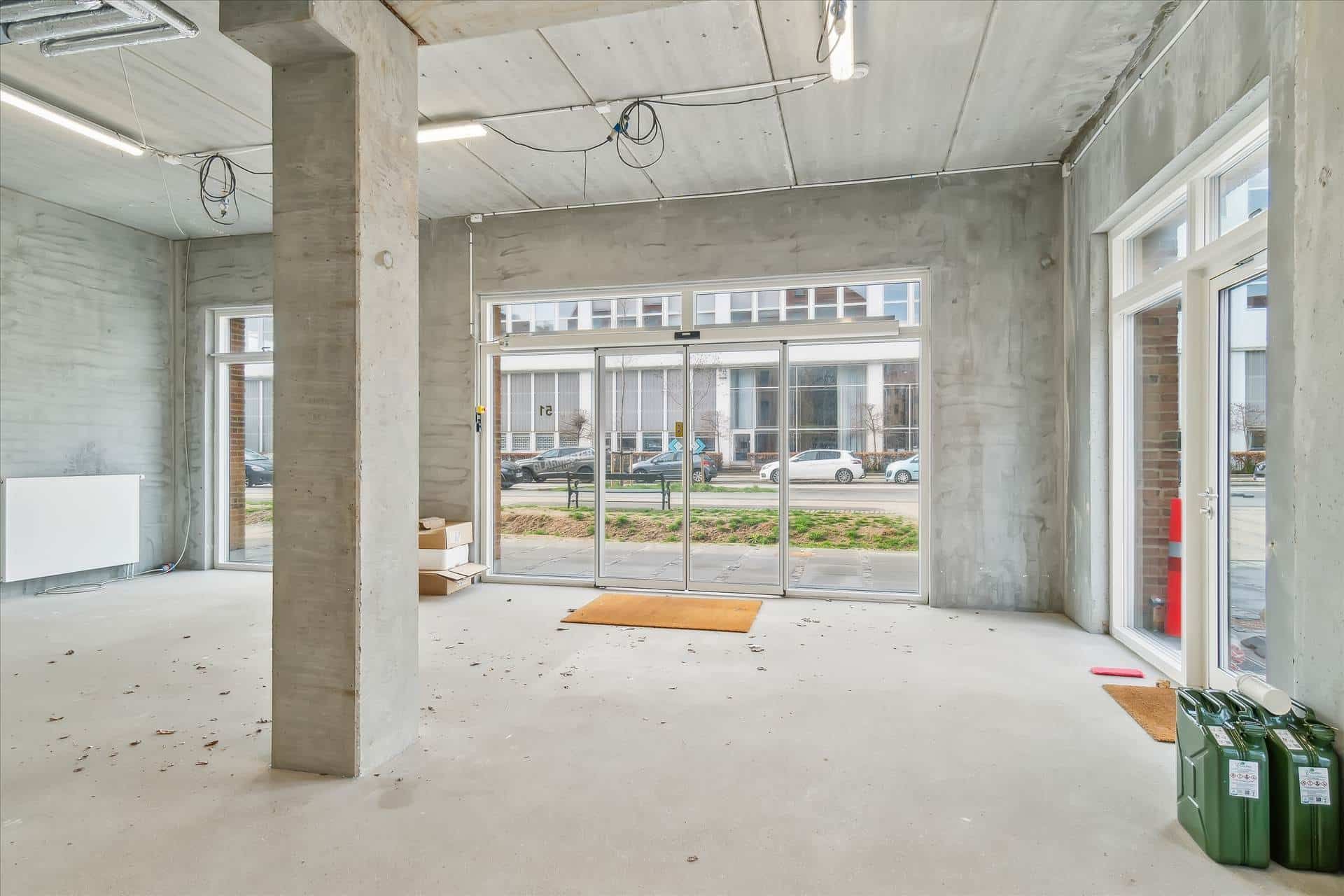 En tom kontorfællesskab med store ruder og tæt ved en parkeringsplads, hvor biler er parkeret. En gul gulvmåtte og to grønne