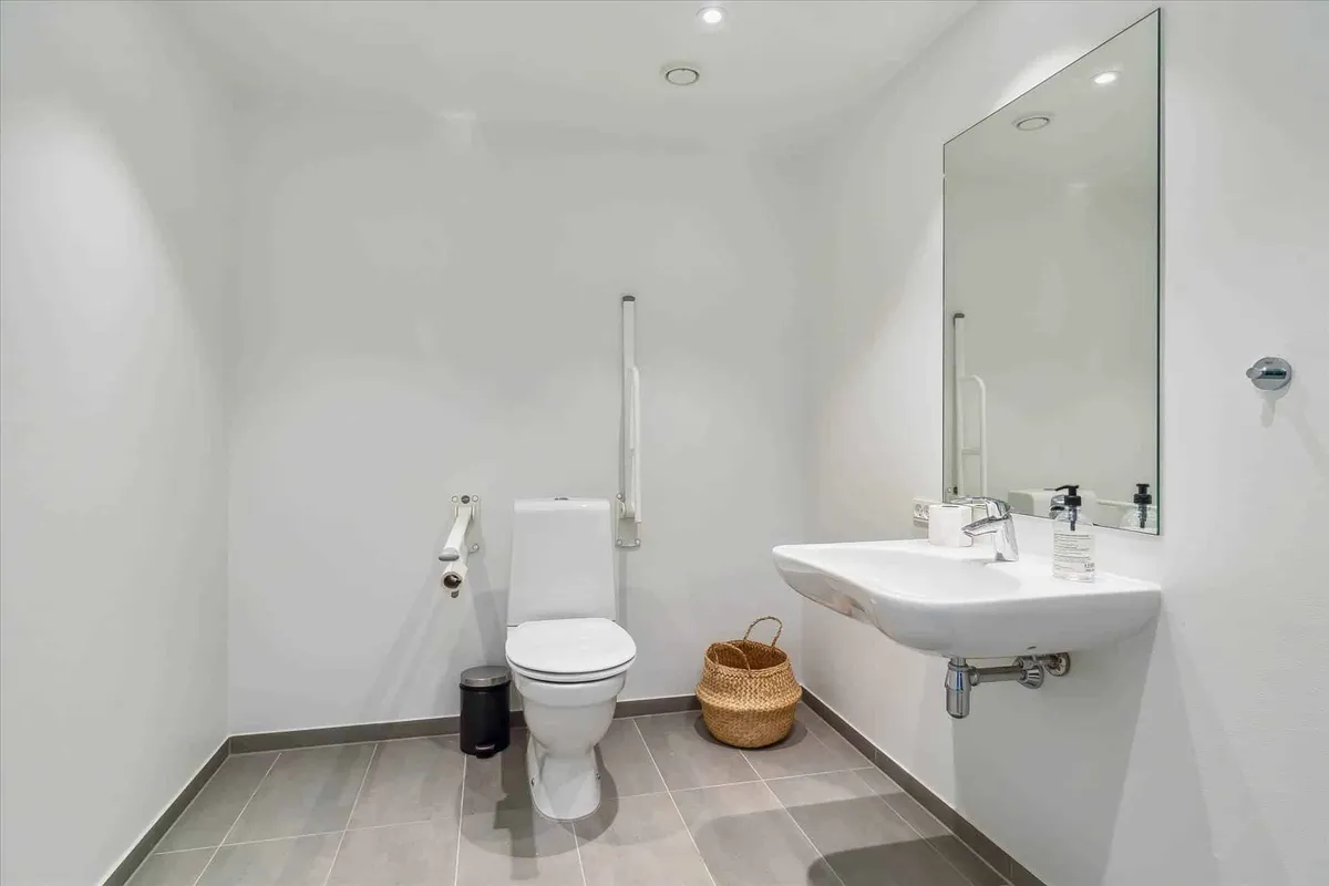 Et lille badeværelse med hvidt toilet, ren hvid vask, smykket med en lille kurv og en spejl med håndtag