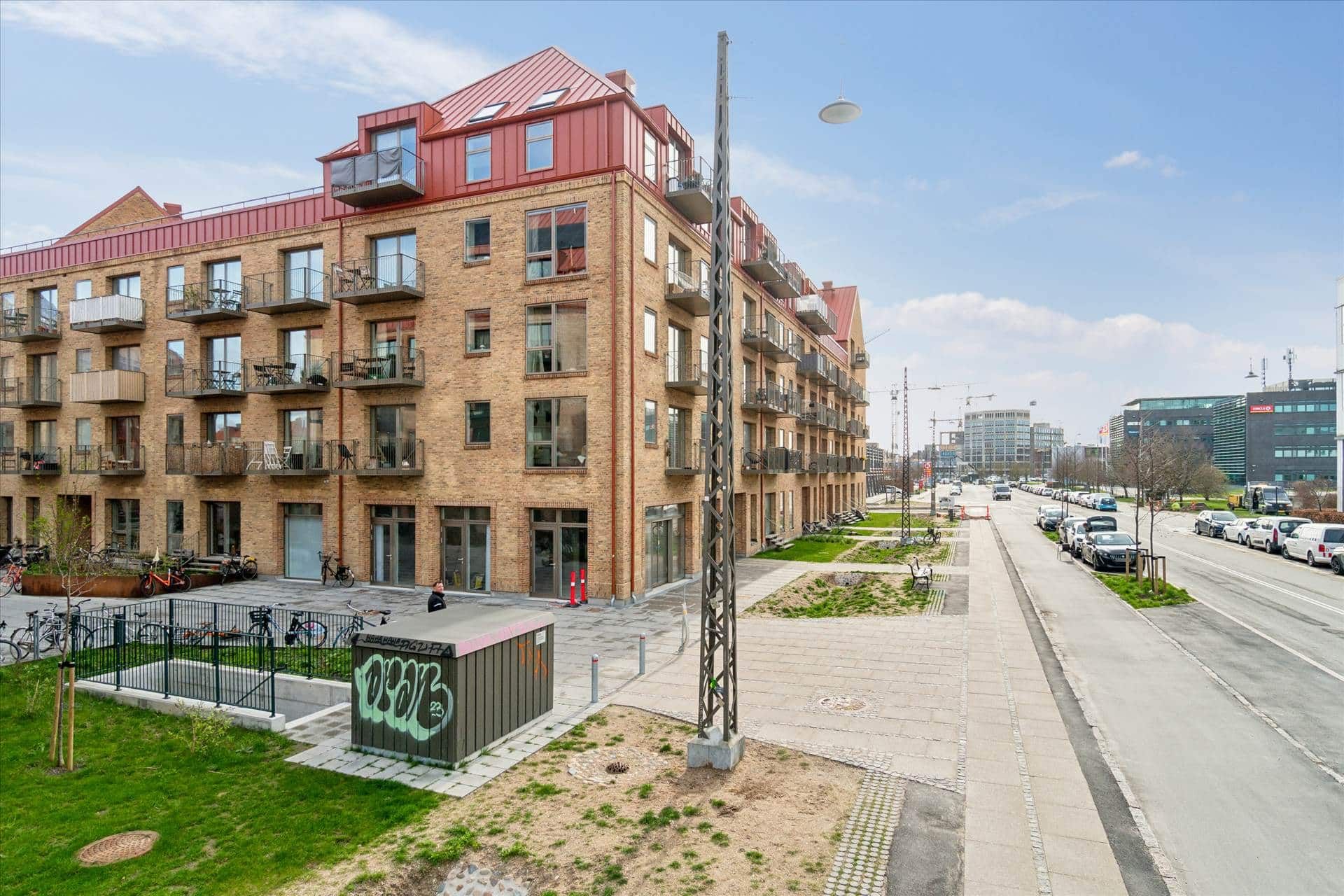 Et moderne bygningsværk med røde tager og balkoner ligger ved siden af en vej med parkerede biler og en gågade med træer og