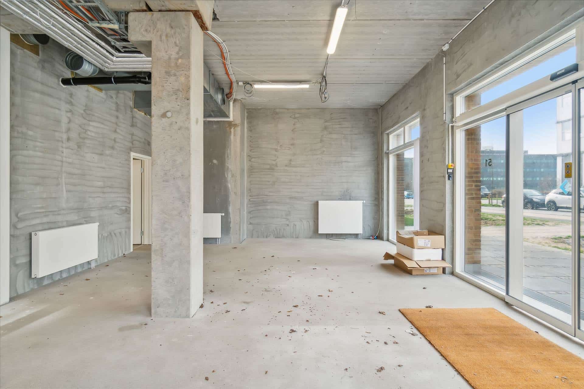 Store glasruder, hvide vægge, hældningsbelægning, radiatorer, trægulve, lagerkartoner, to tæt parkerede biler og en