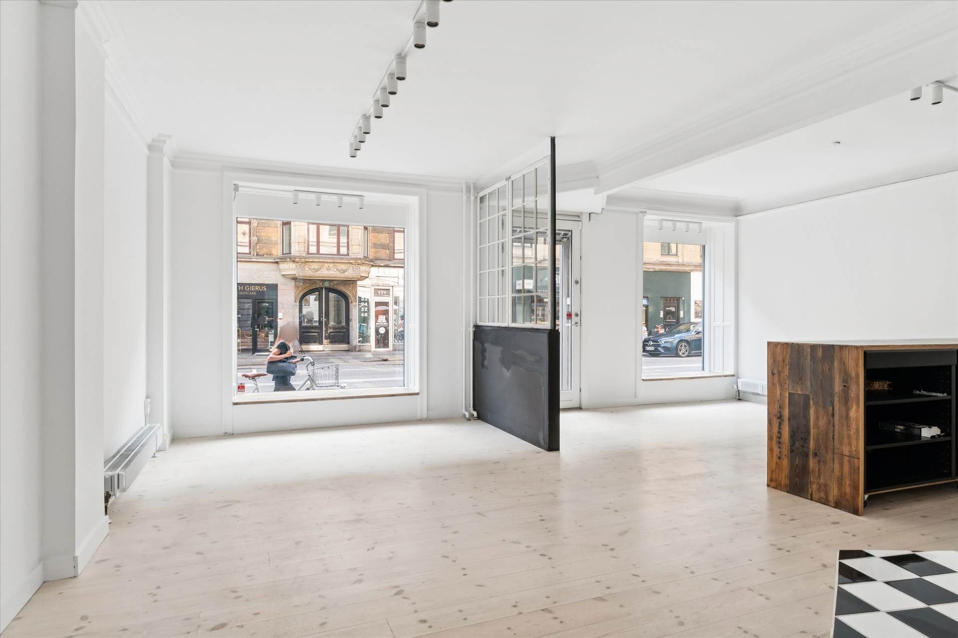 Store vinduer og minimalistisk indretning med hvide vægge, parketgulve og lysekroner i loftet. Sorte møbler og træhylde med