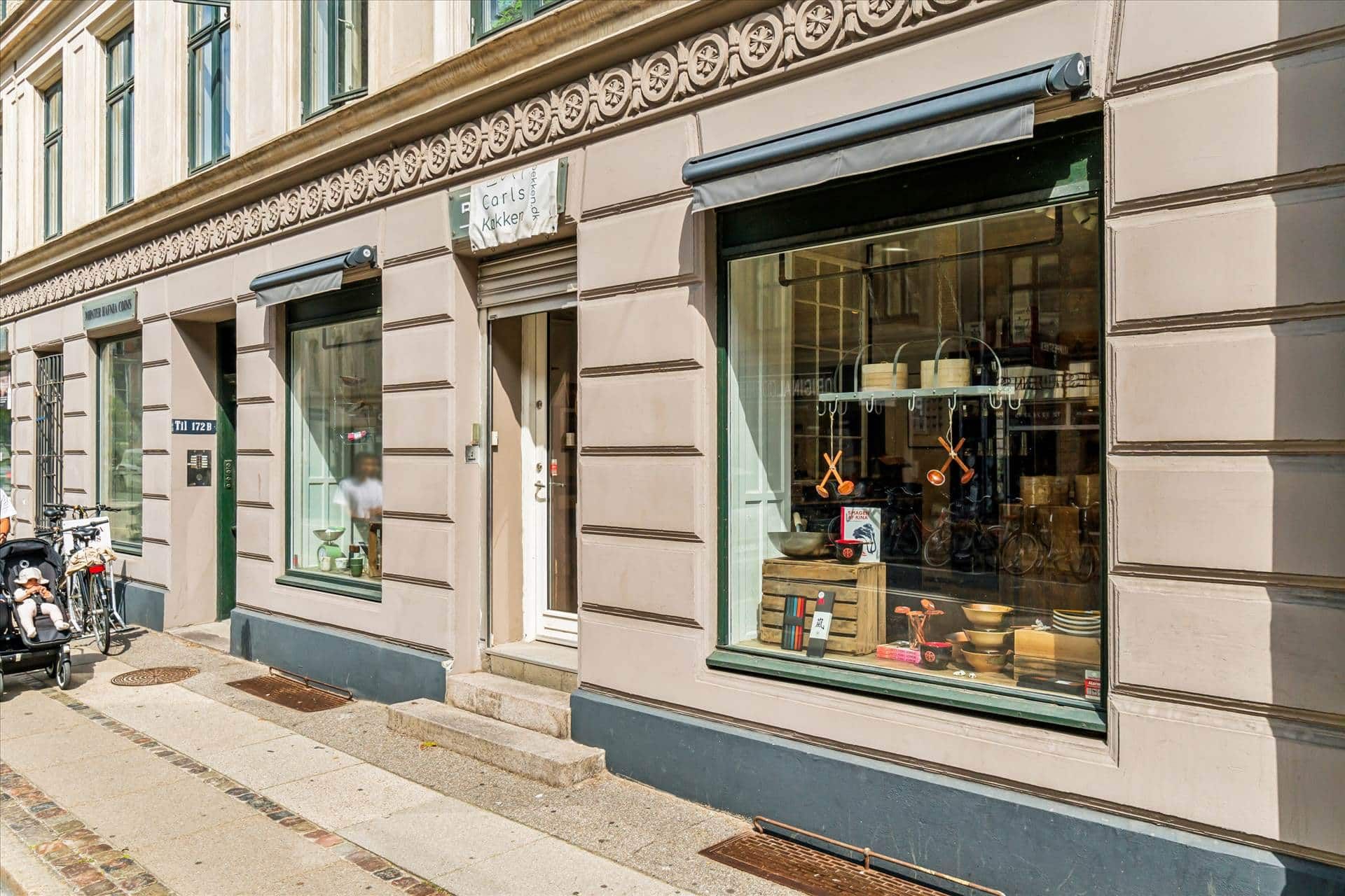 Butik med store vinduer og udskæringer i gul puds, hvor der står Carls Køkken, og udsigt til en vogn med barn og en