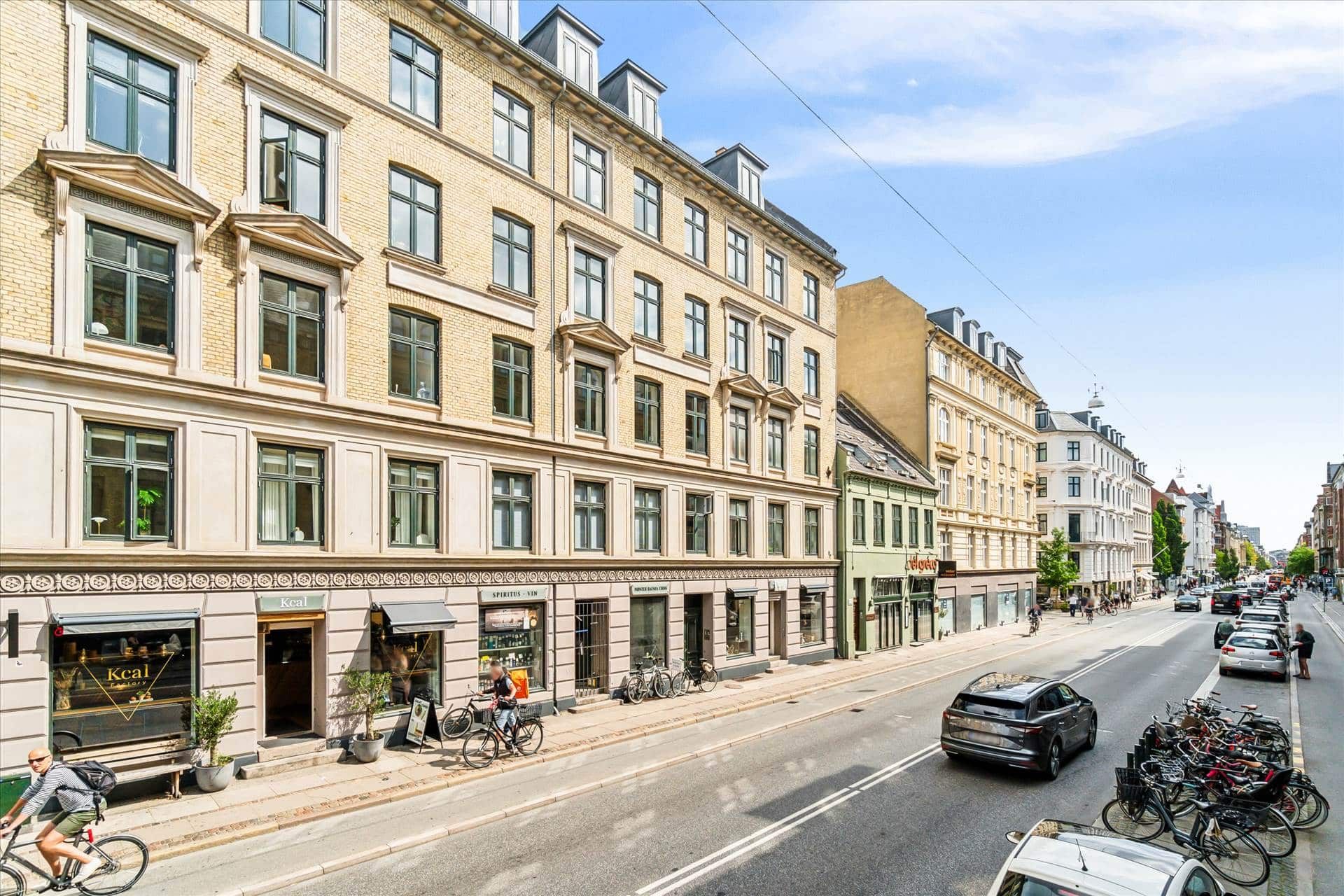 Biler og cykler kører ad gade med bygninger og grønne plantager i baggrunden