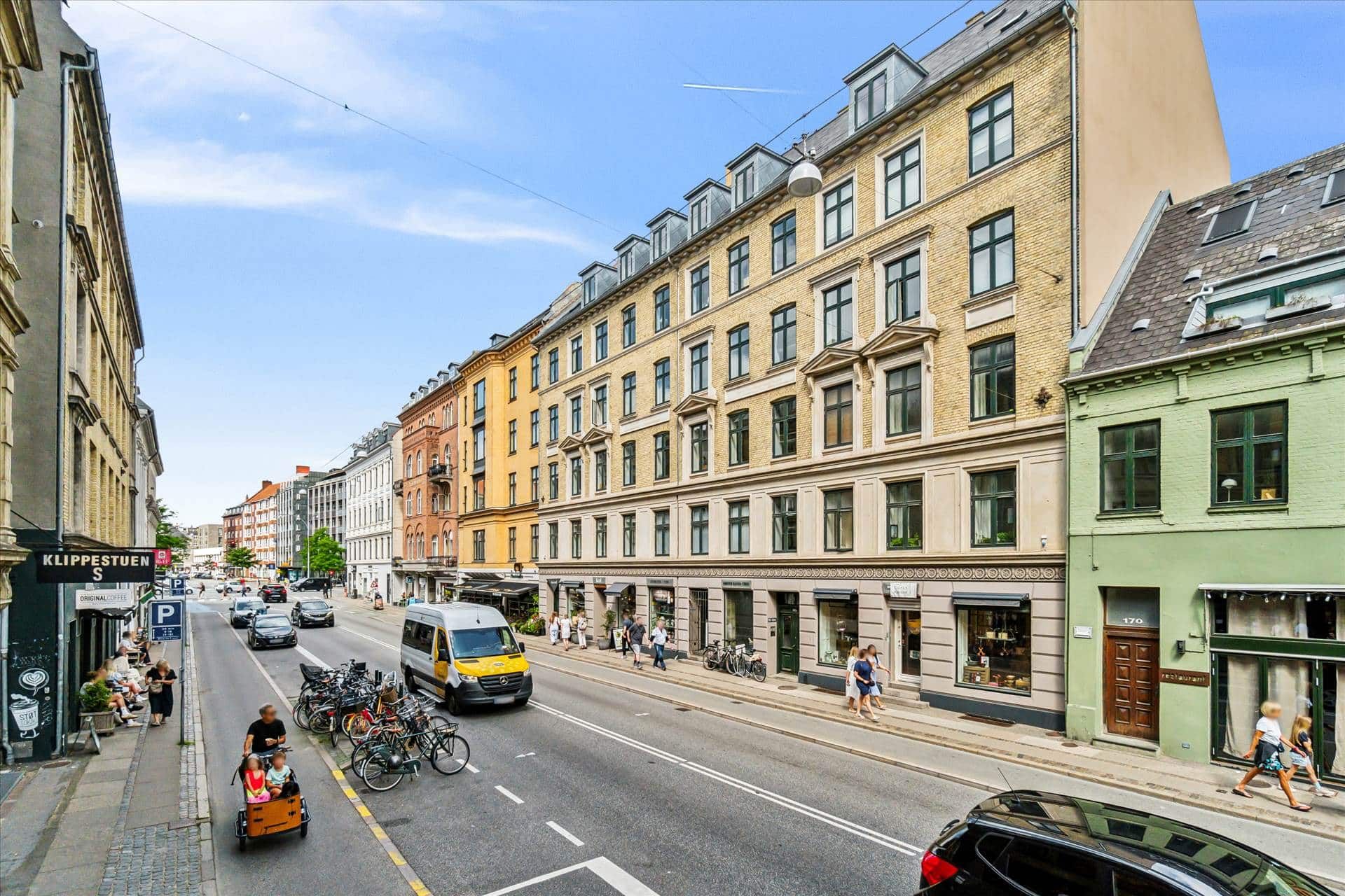 En gågade med biler og cykler med mennesker, der går og går i forgrunden, og en vandbus og en bil, der kører i baggrunden.