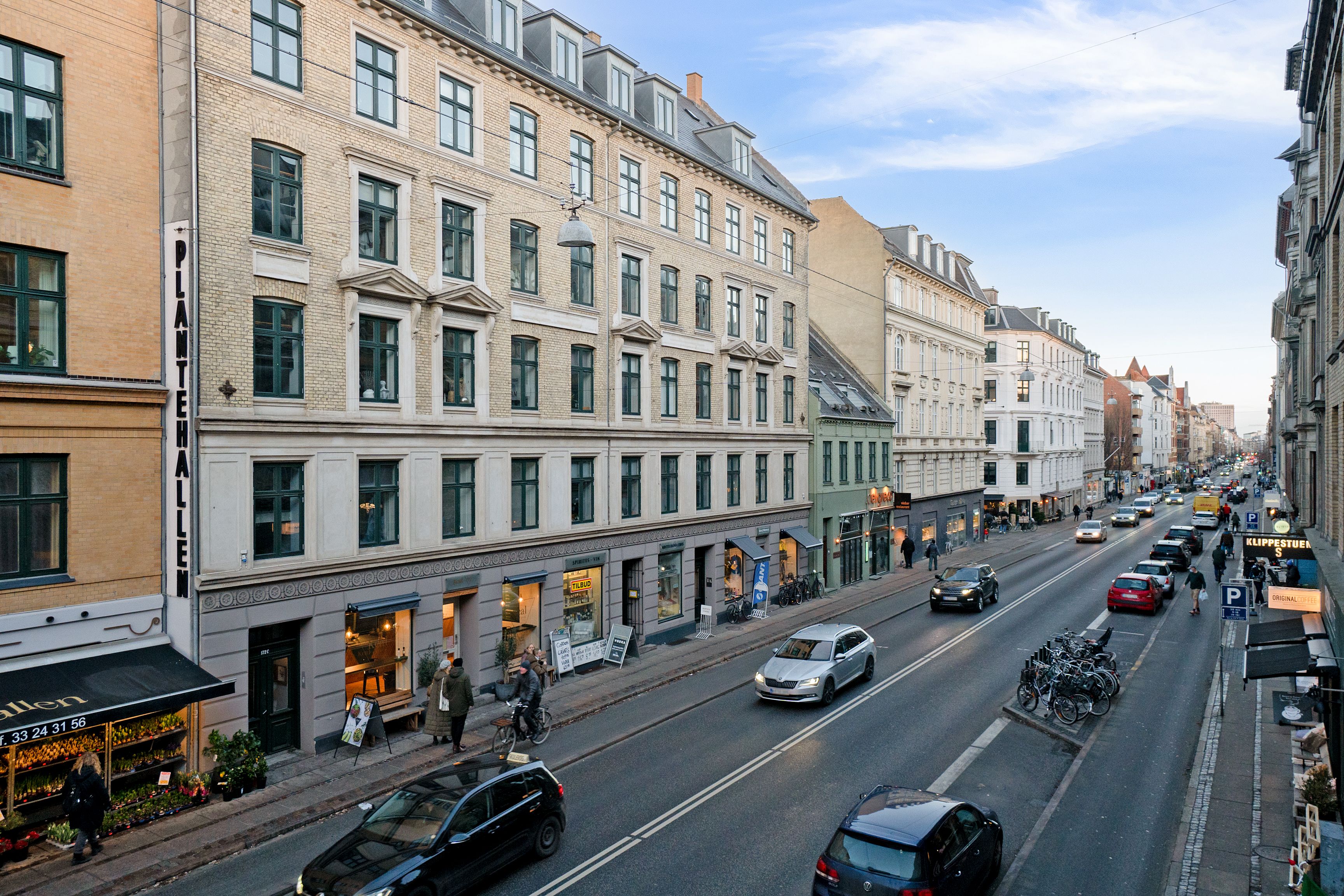 Mennesker går på gågaden og kørebilerne kører langs den. Bygningerne med vinduer og butikker står langs vejen.