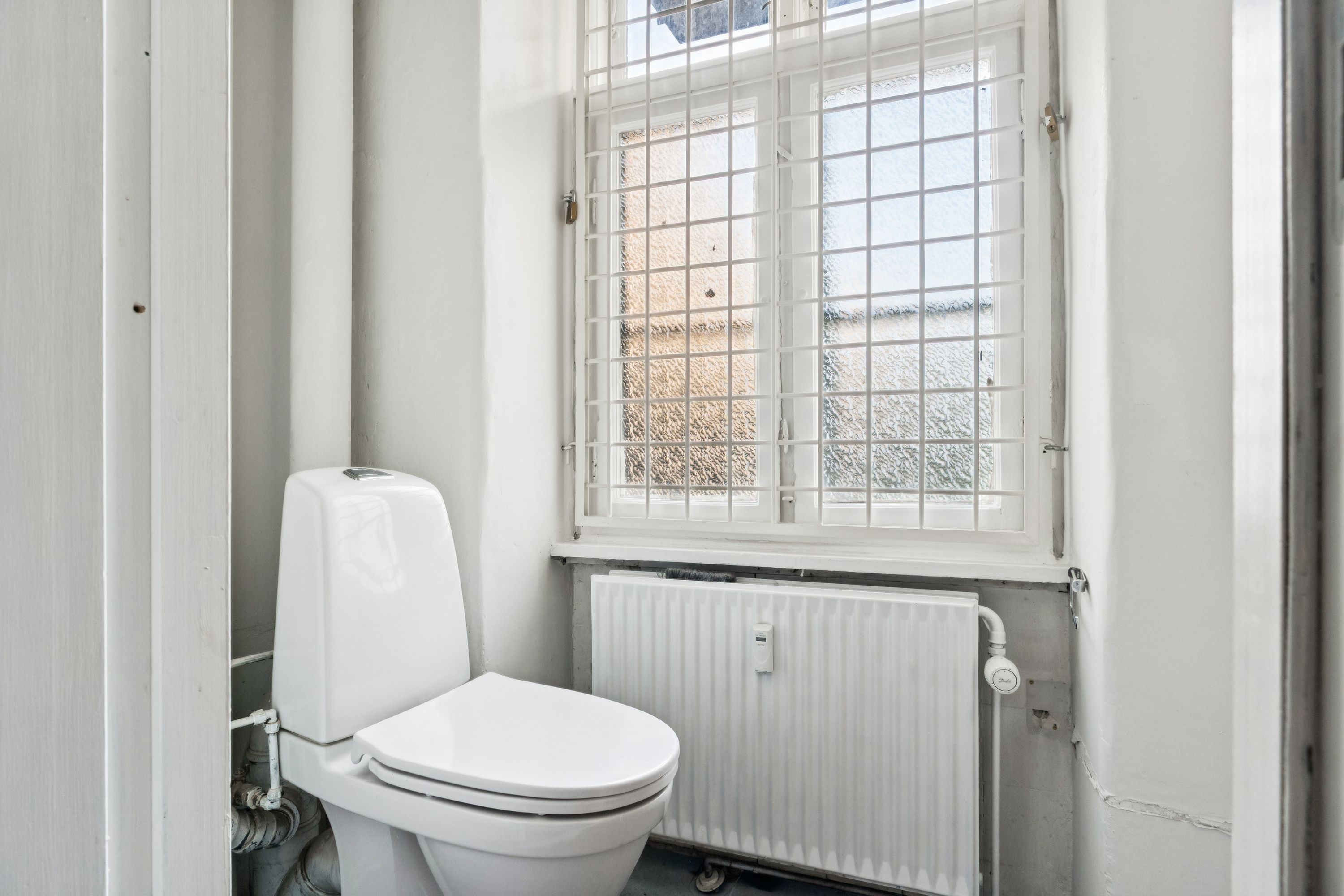 Moderne toilet i lille badeværelse med hvid ventileret vinduesruder og lysegrå radiator. Lysegulv og hvide vægge.
