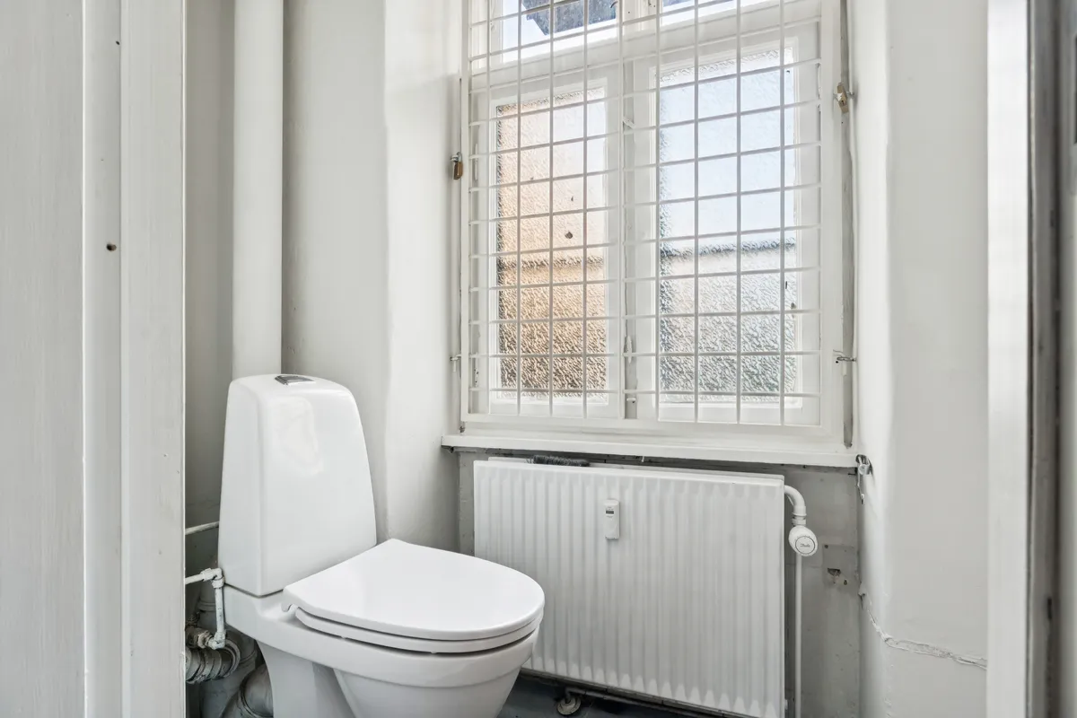 Moderne toilet i lille badeværelse med hvid ventileret vinduesruder og lysegrå radiator. Lysegulv og hvide vægge.