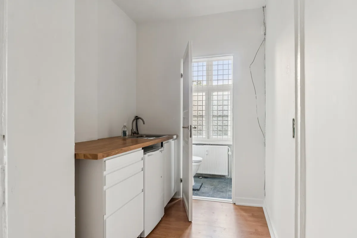 Billede af et lukket hjørnekøkken med hvide vægge og trægulv, et hjørne med håndvask og en radiator i det nærliggende badeværelse