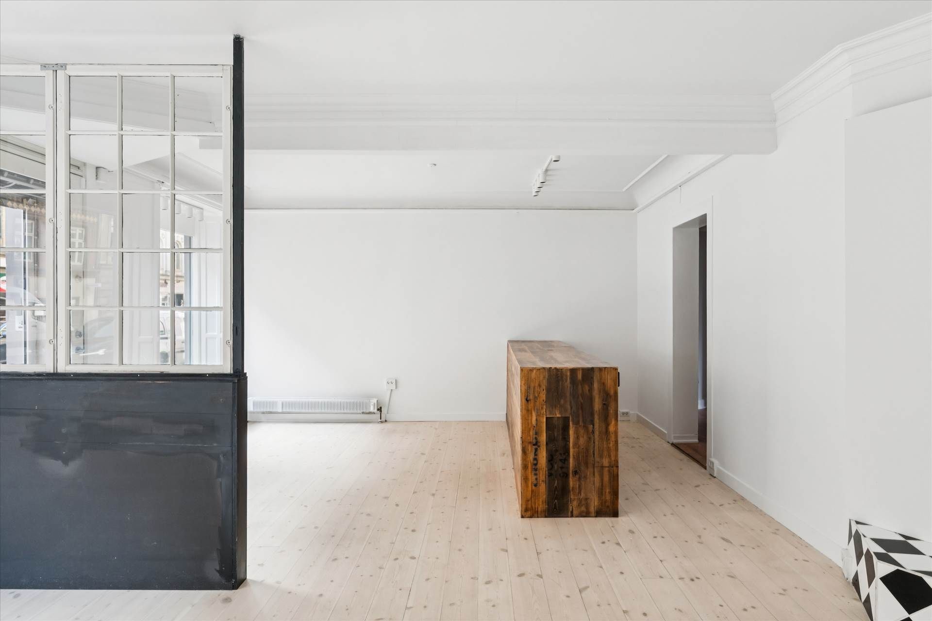 En tom rum med hvide vægge, lyse gulv og mørkt sidebord, med et stående træskab og åbne ruder
