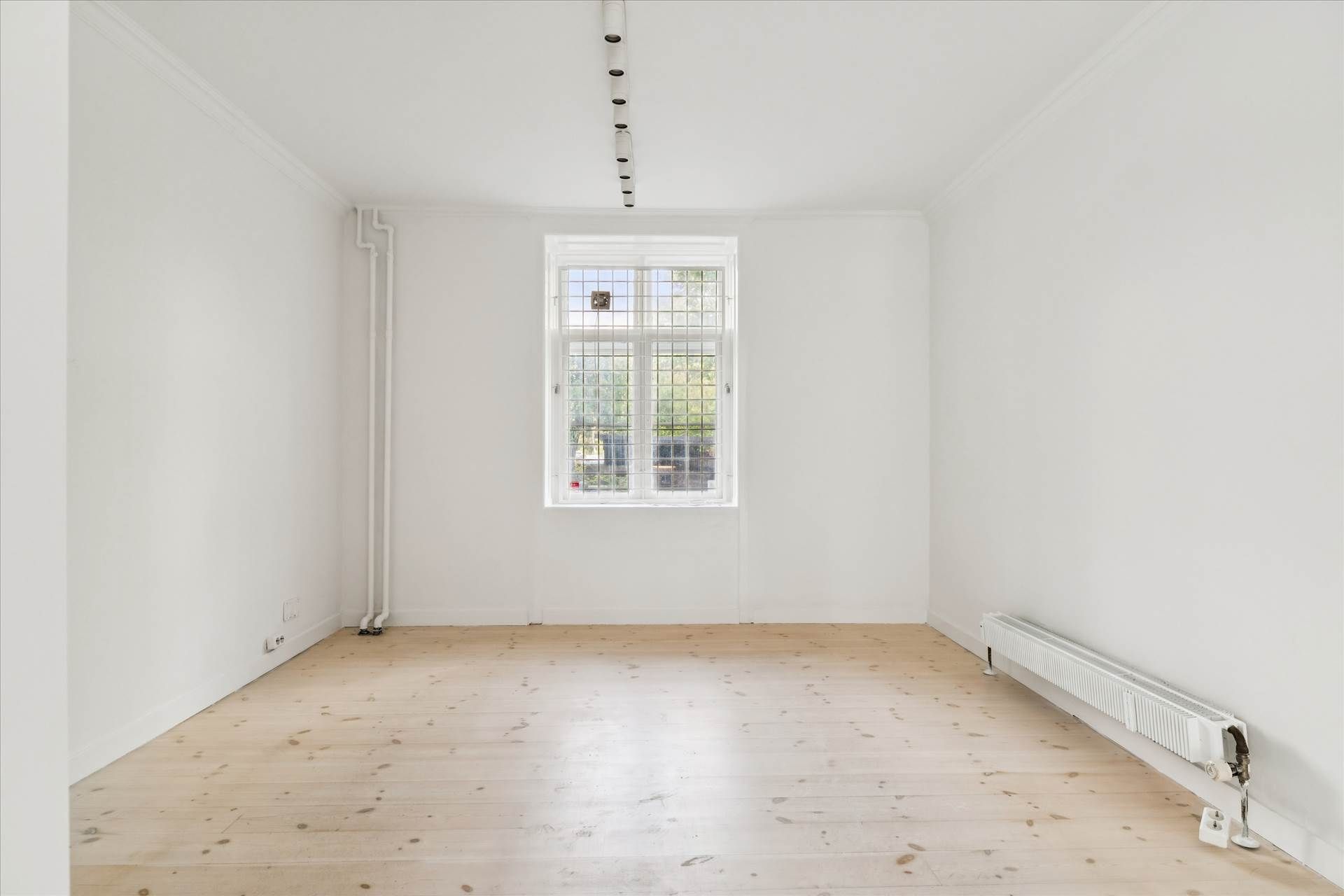 Et tomt værelse med hvide vægge, lysegrå gulvbelægning og en stor enkeltvindue. En radiator og en række lyspærer er monteret