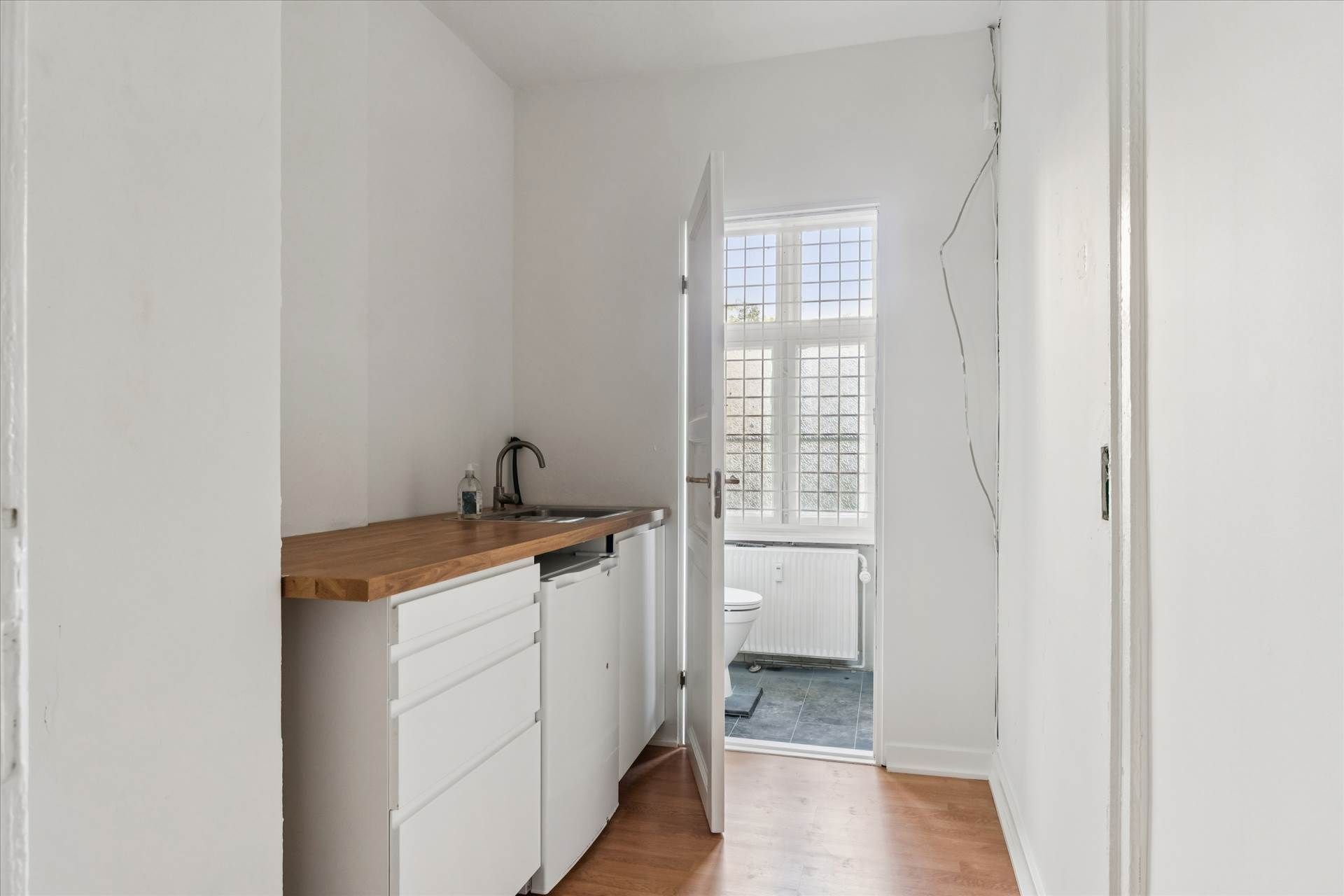 En tom lyse stue med hvide vægge og trægulv og et lille køkken med en hætte over vasken og en toilet i siden.