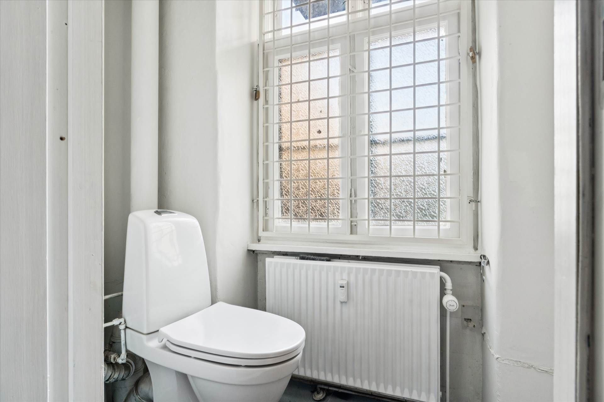 Et badeværelse med et hvidt toilet, et radiator og et vindue med en metalgitter og glaspaneler