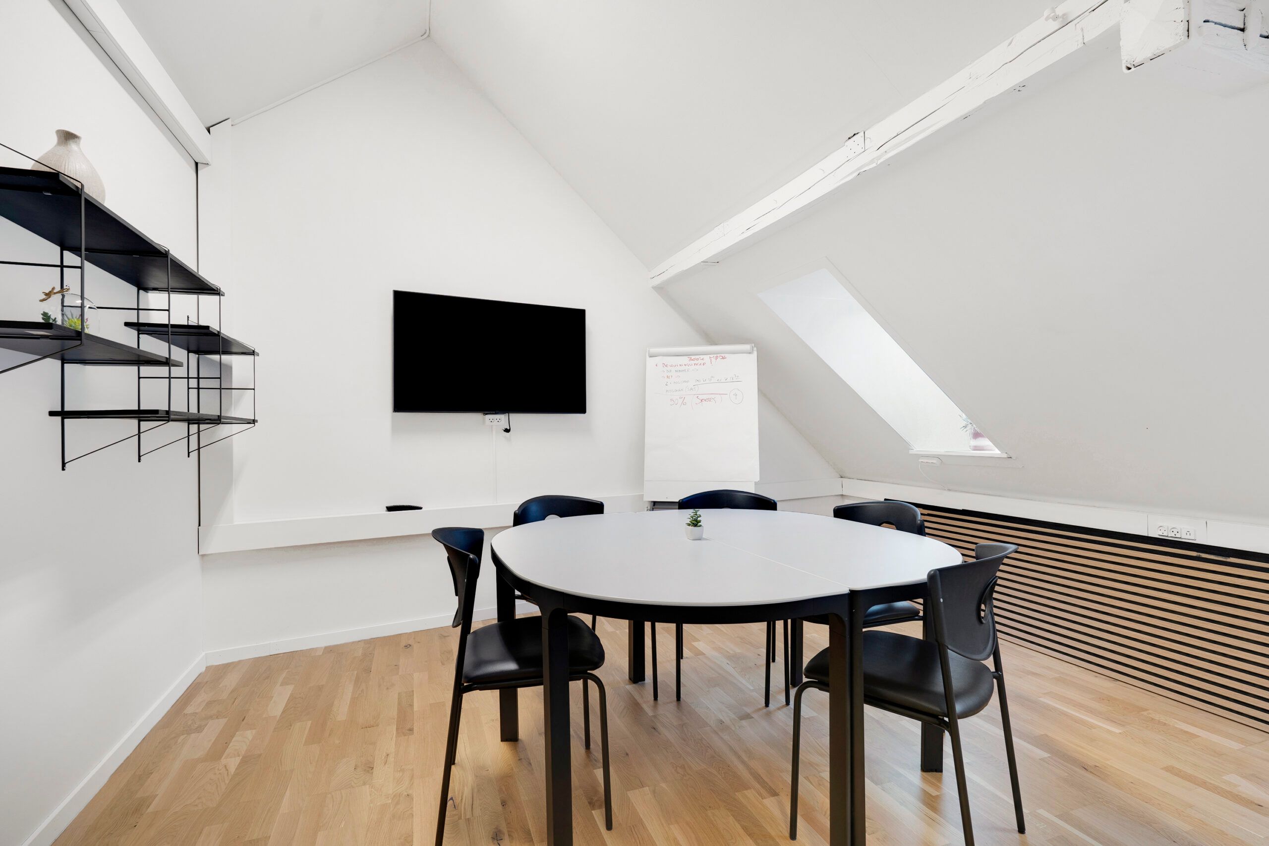 Kontor med højt loft og lyse gulve. Rundt bord med flere stole rundt. En tv og et hvidt papir med tekst skråt på væggen.