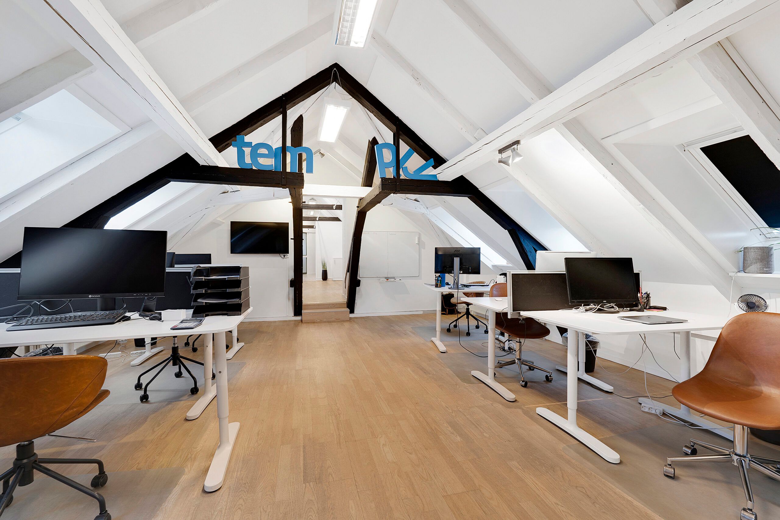 Kontorlokaler med højloftede rum og moderne møbler, skriveborde og skærme, oplyst af lyse loftslamper og lyse gulvbelægning