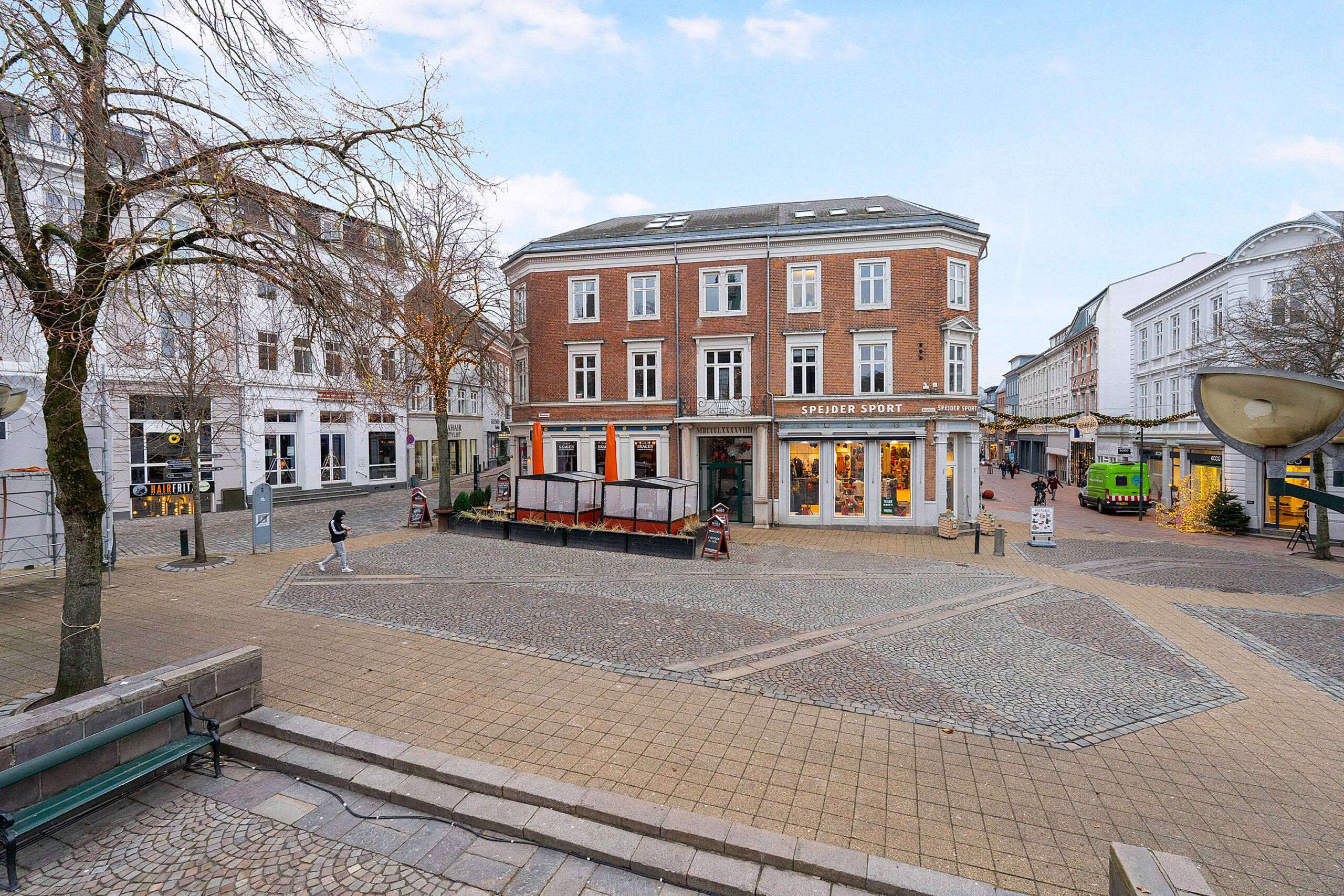 Bred og gågade med kontorer og butikker, en person vandrer og der er flere gående mennesker i baggrunden