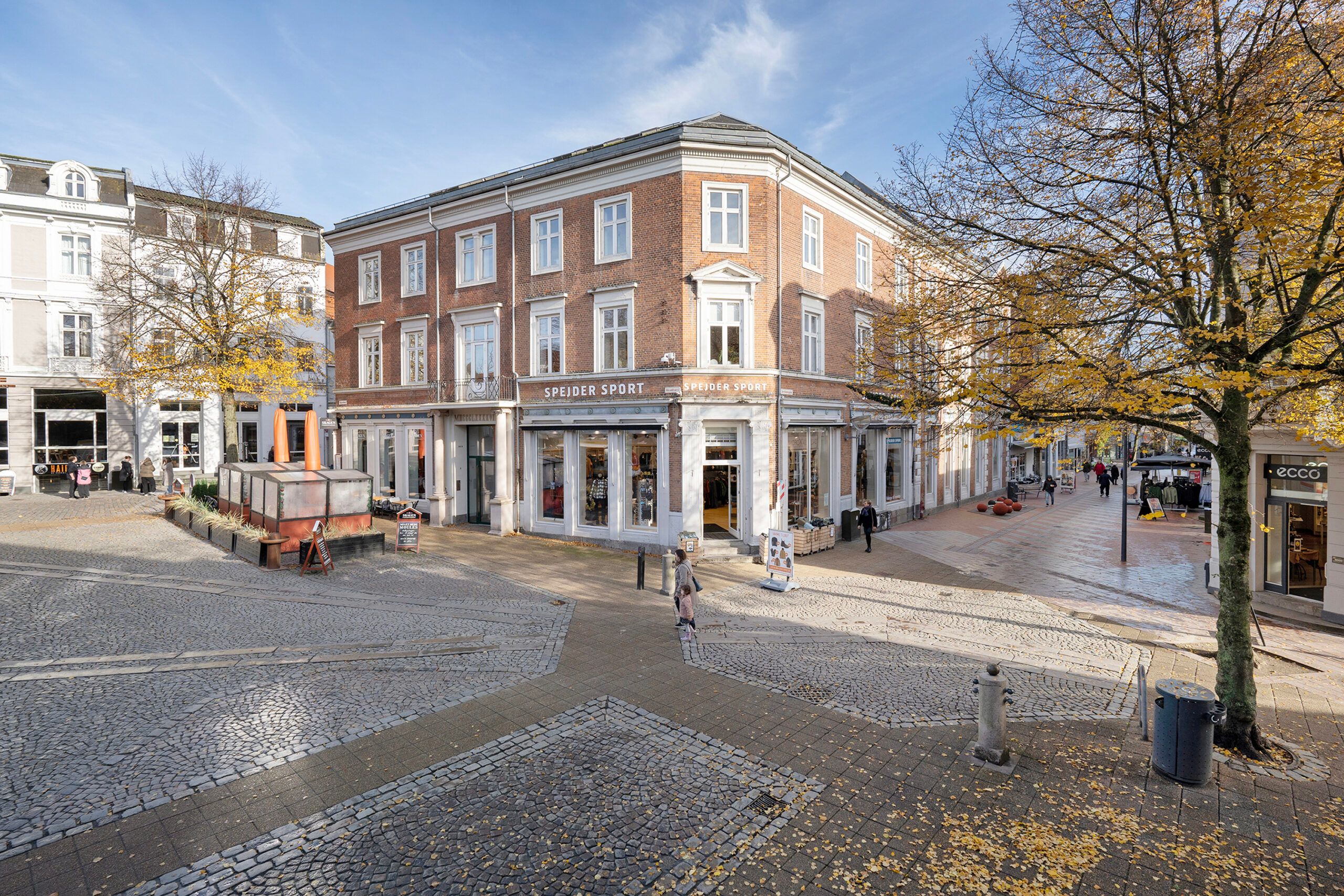 Historisk bygning med butikker og kontorer i Koldings centrum, hvor en person går forbi et skilt med teksten "Speidier Sport"