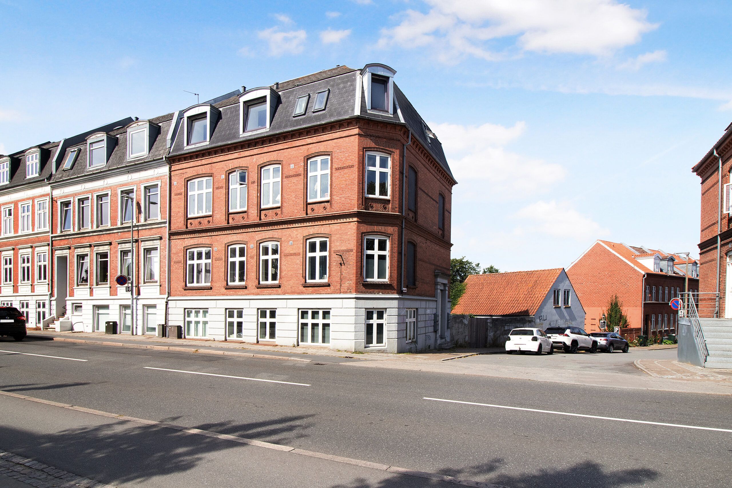 Tre biler parkeret i garagen og en stor bygning med røde mursten og hvide vindueskarme