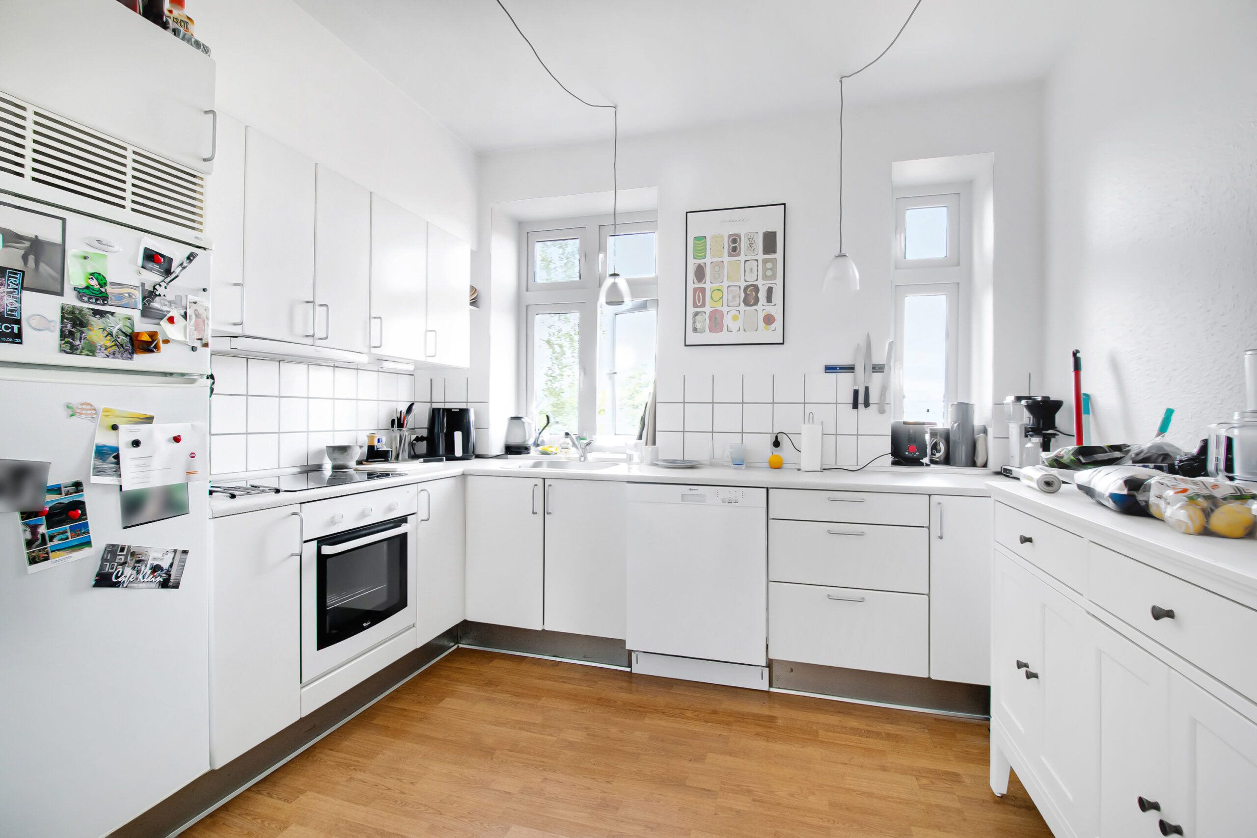 Rent og ryddet køkken med hvide vægge og trægulv, hvidt køkken med hvide skabe og møbler, hvide vægpaneler og tre vinduer