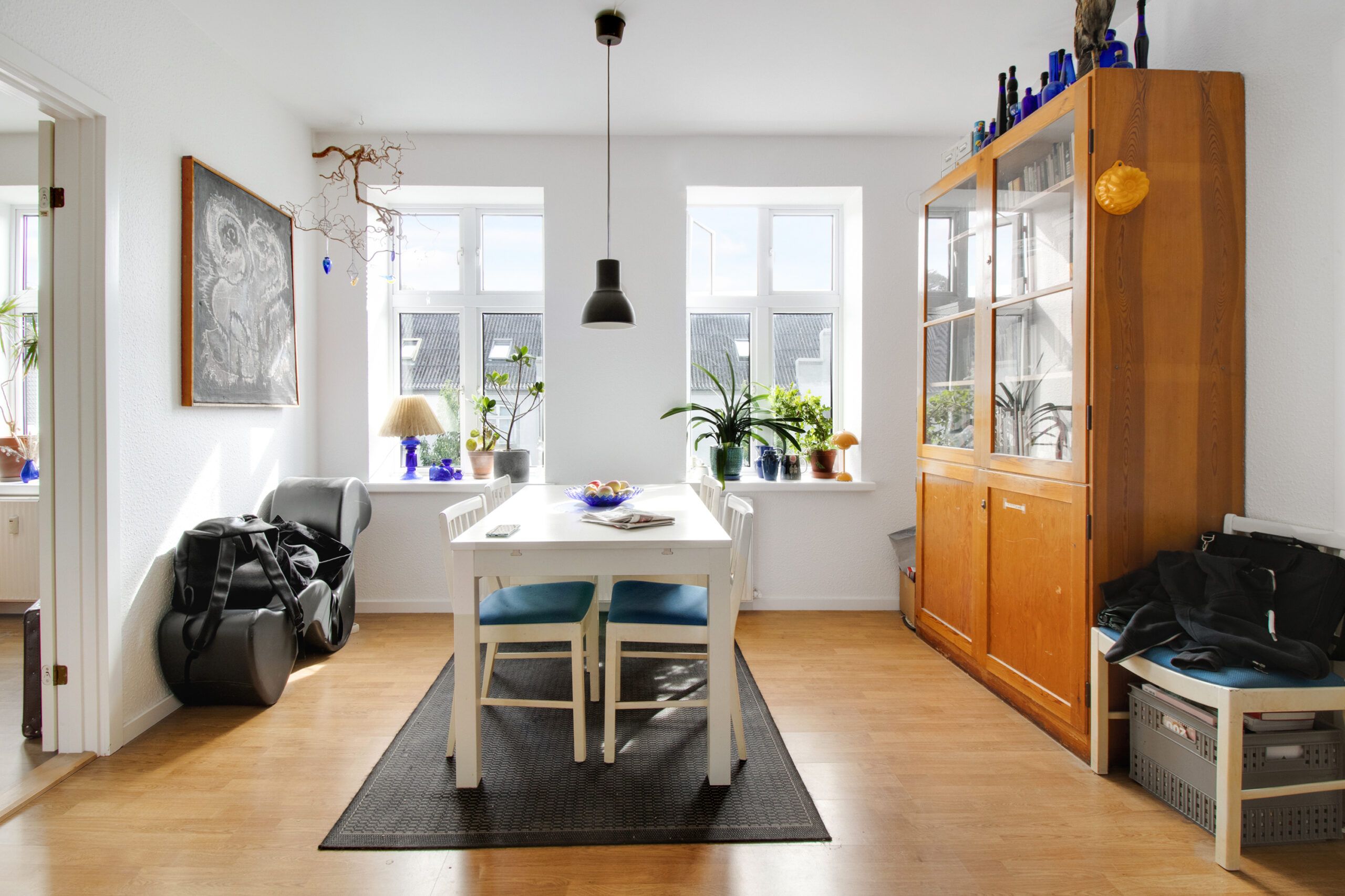Foran et vindue er der et hvidt spisebord med fire blå stolen og en lysebrun gulvmåtte