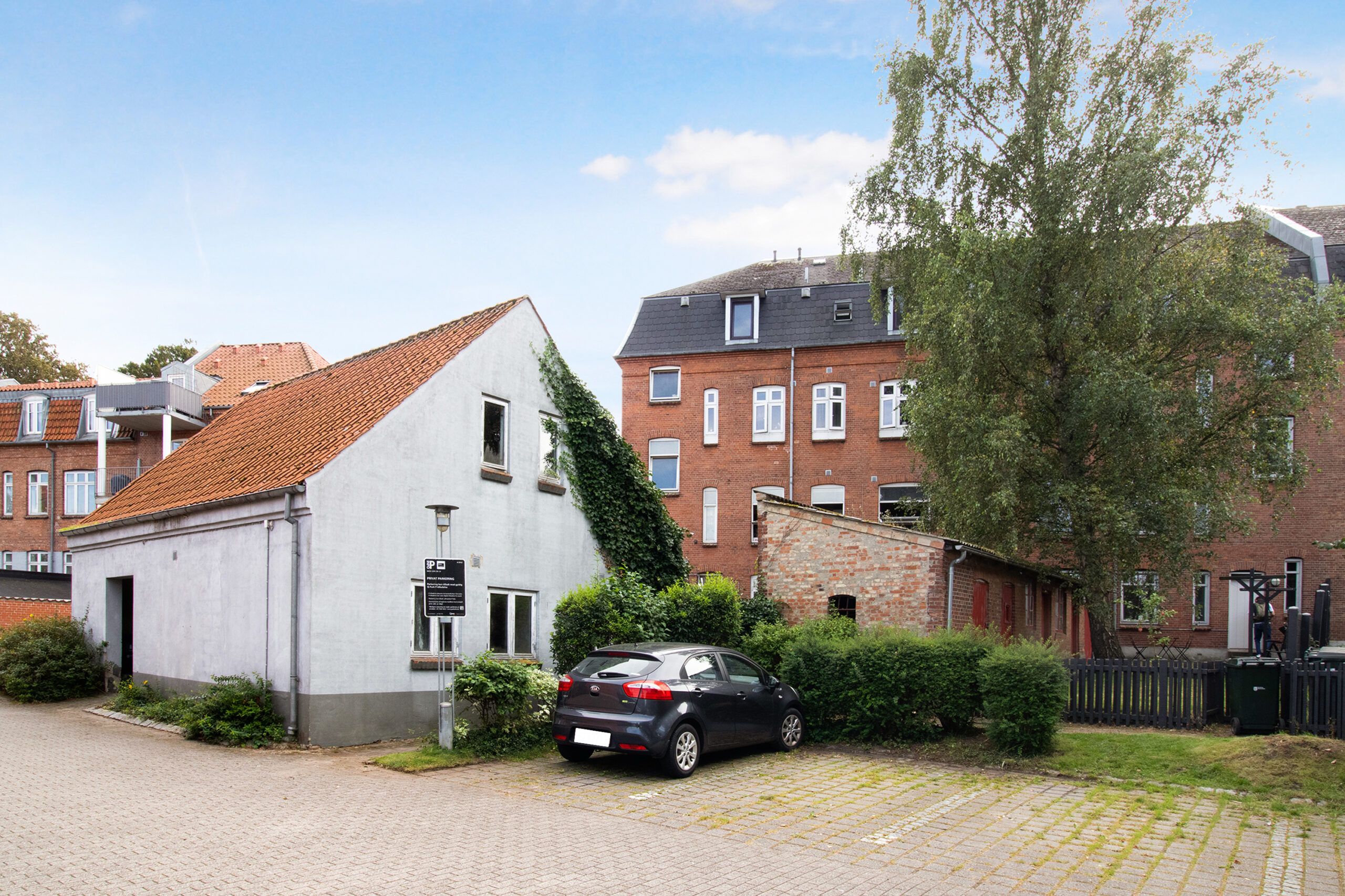 En gul husbygning med et brunt tag og grønne planter samt en gul bil parkeret foran.