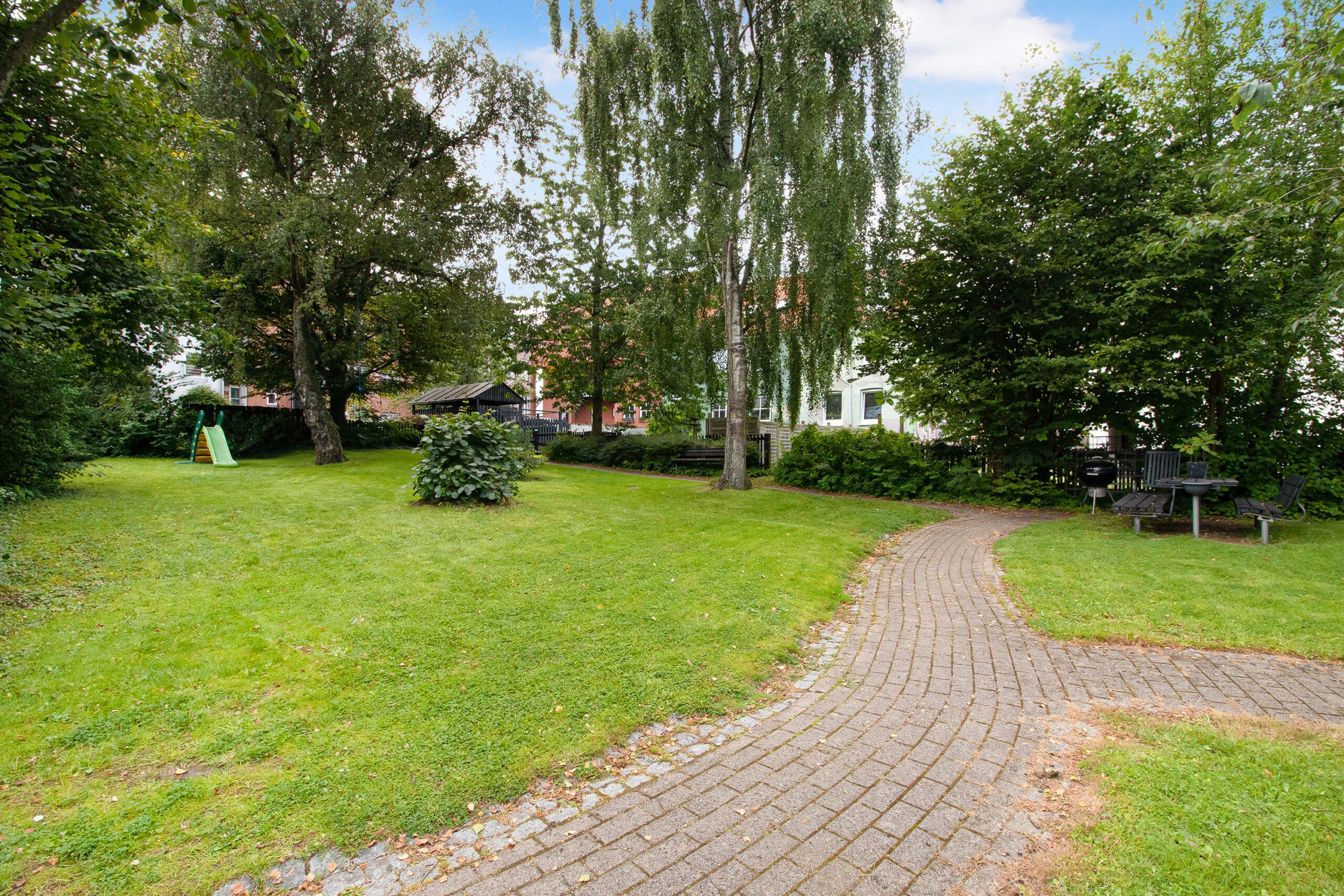 En park med stier og græsplæne, plantager, en grøn børnespildække og grønne planter med en husbygning i baggrunden