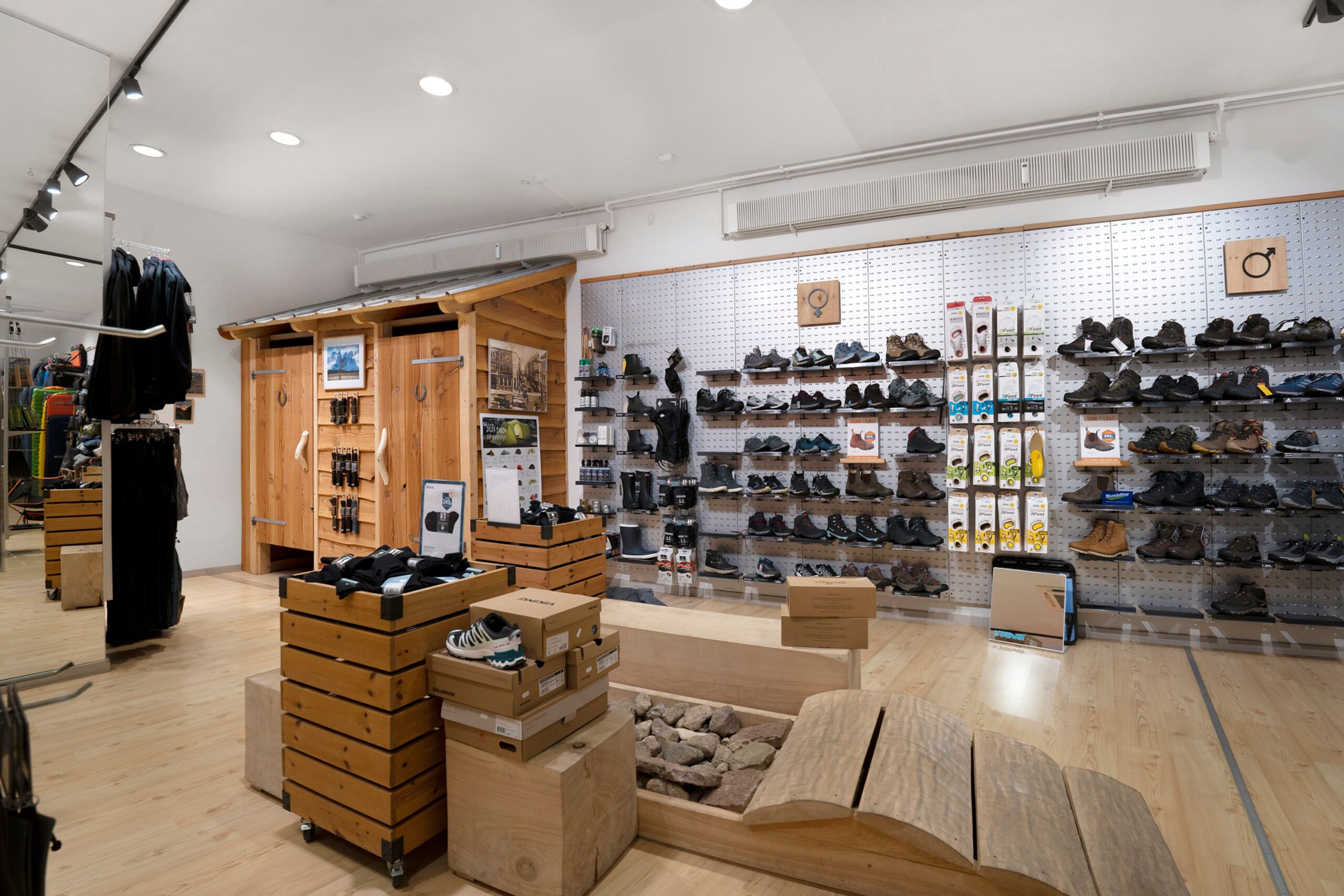 Indvendigt hos en butik med træhylder, sko samt tøj og trækasser med tøj og sko. Høje lofter med lysekroner og ventiler samt