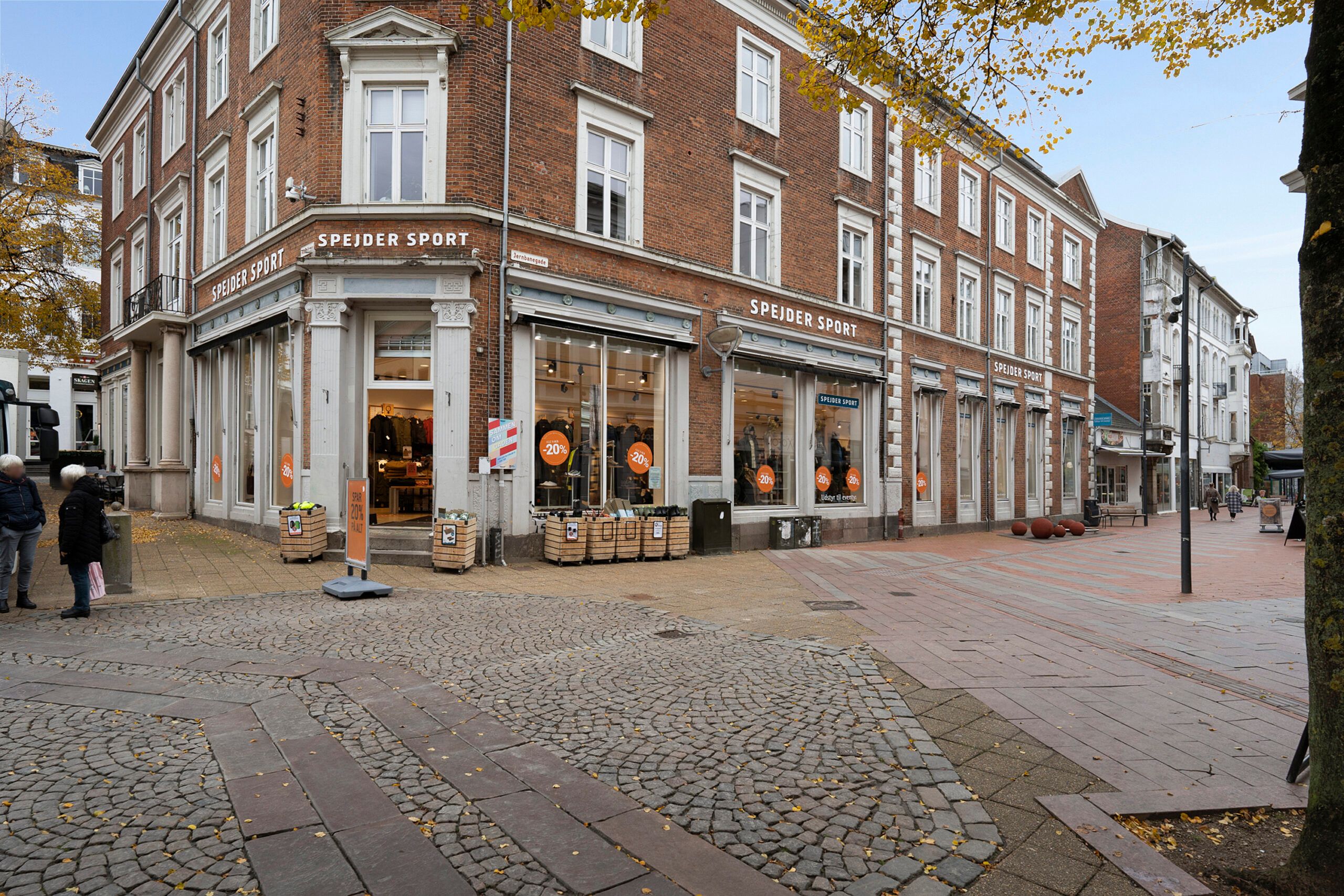 Bred gågade med sælgere og gående forbi butikker og rådhuset i baggrunden med spejder sport og spar udsalg