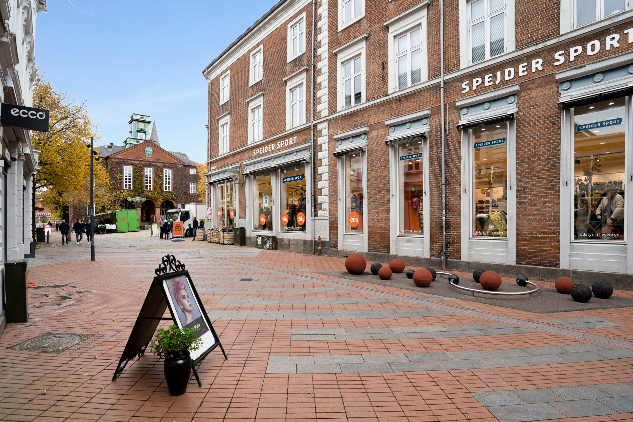 Lejemål i historisk ejendom, med udsigt over rådhuset, butiksfronten for Spøjder Sport, og folk vandrer på gaden
