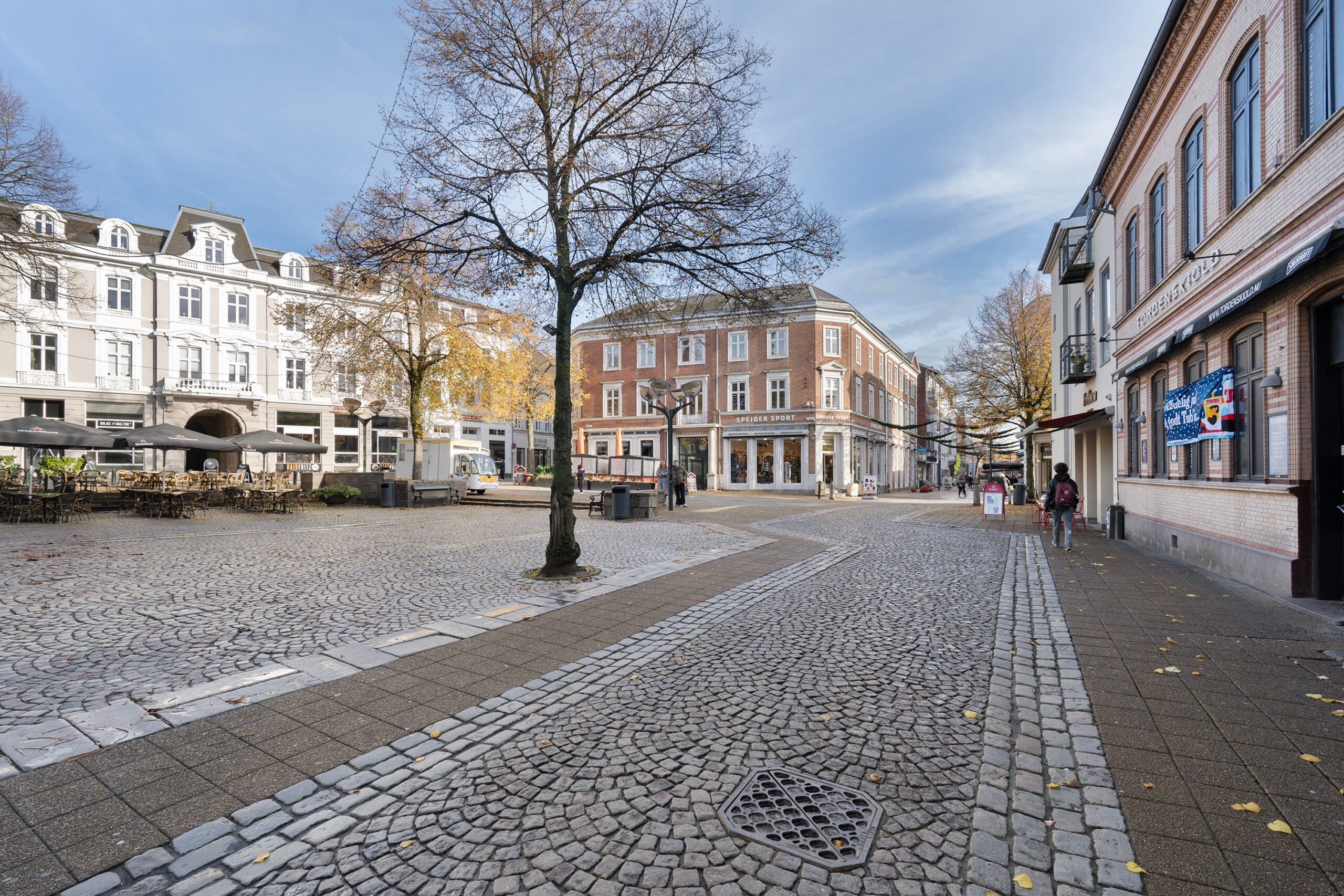 En fredelig gågade med træer og pavilloner, der vender ud mod et stort, rødt bygningskompleks. Her er flere mennesker på