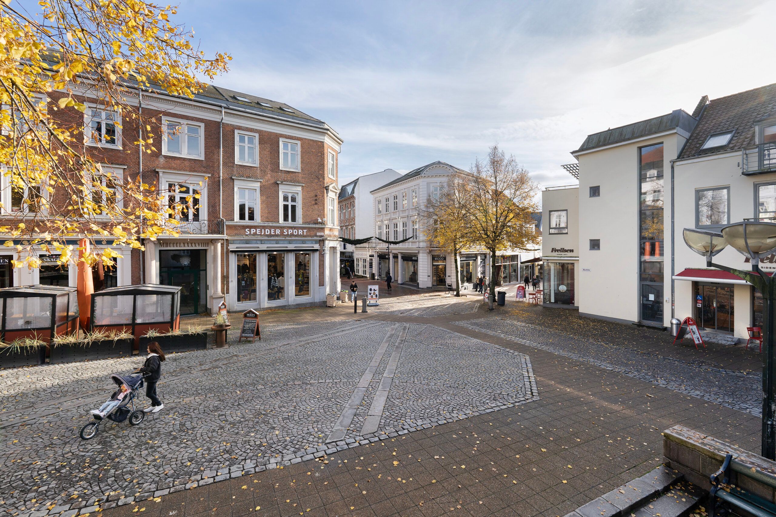 En kvinde med barnevogn går i gågaden med gamle bygninger og en spænder af folk, der går forbi butikker med tekster som