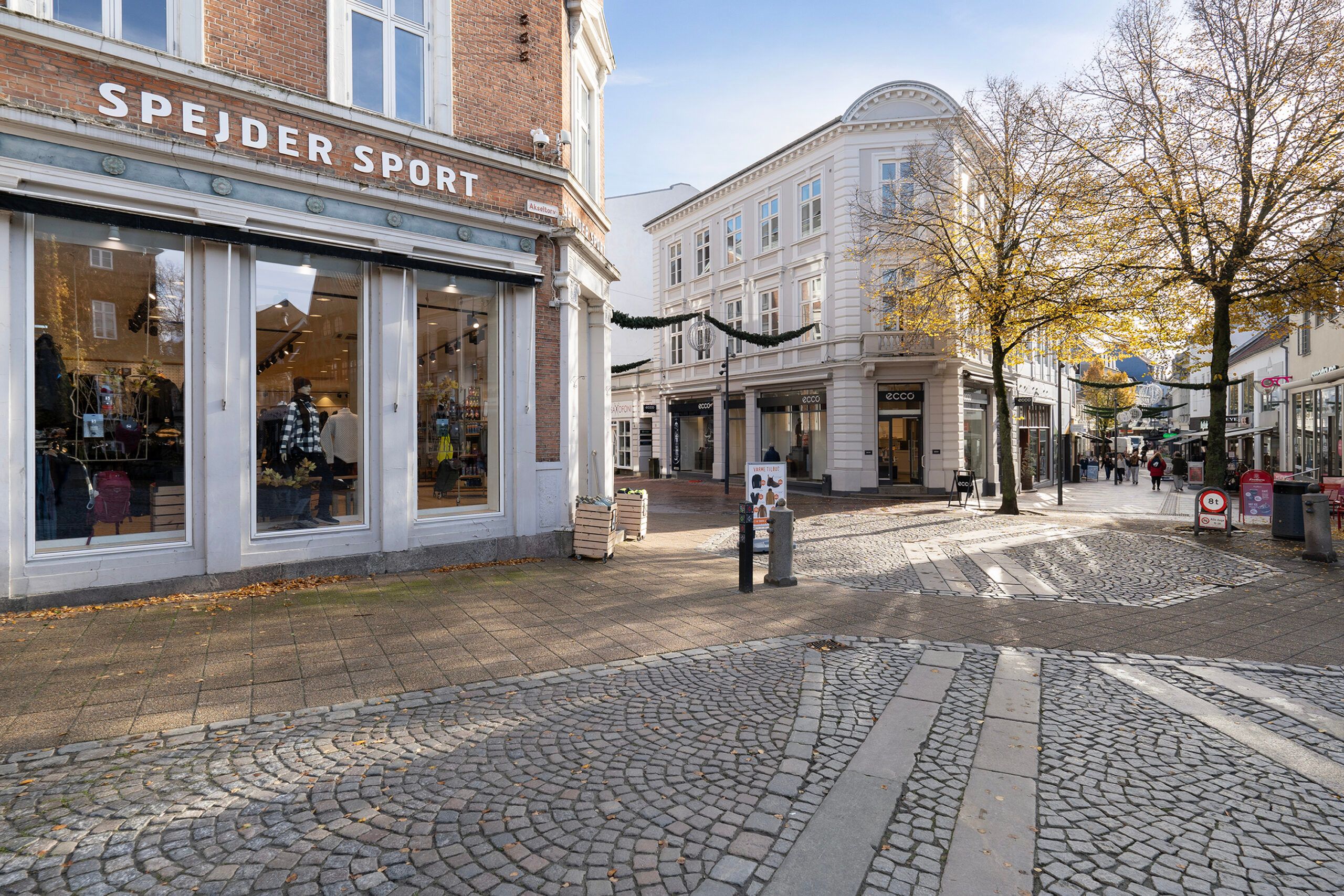 Et stort sportforretning, Spejder Sport, placeret på en bred gågade med træer og folk, der går langs siderne.