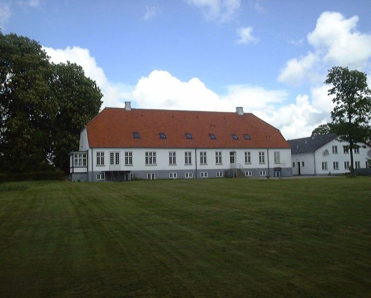 Et stort hvidt hus med rød tagdækning står på en stor græsplæne med mange træer i baggrunden.