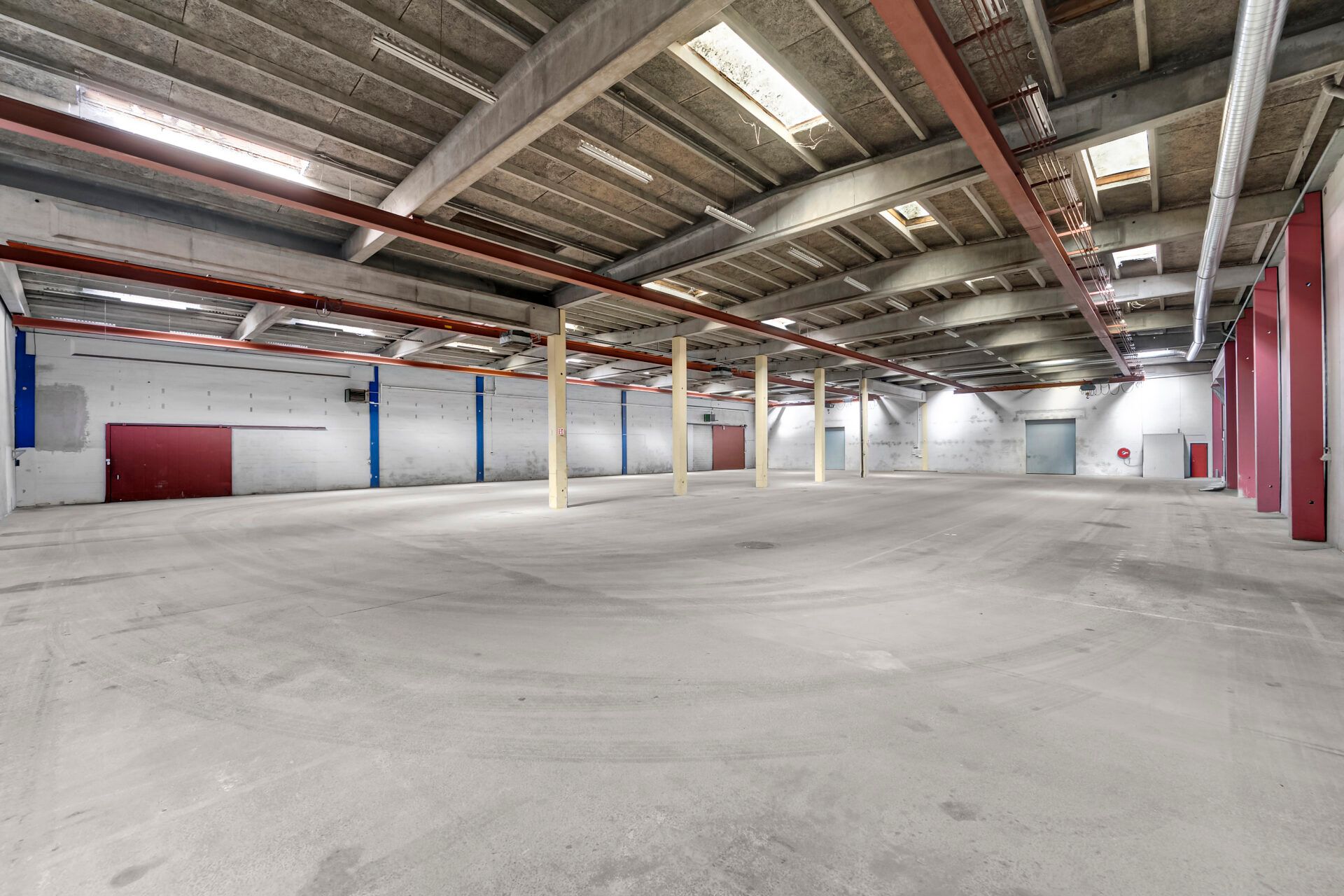 Stort, tomt lager med røde døre og loftsventilationer. Betongulve og røde støtter. Ingen møbler eller mennesker er synlige
