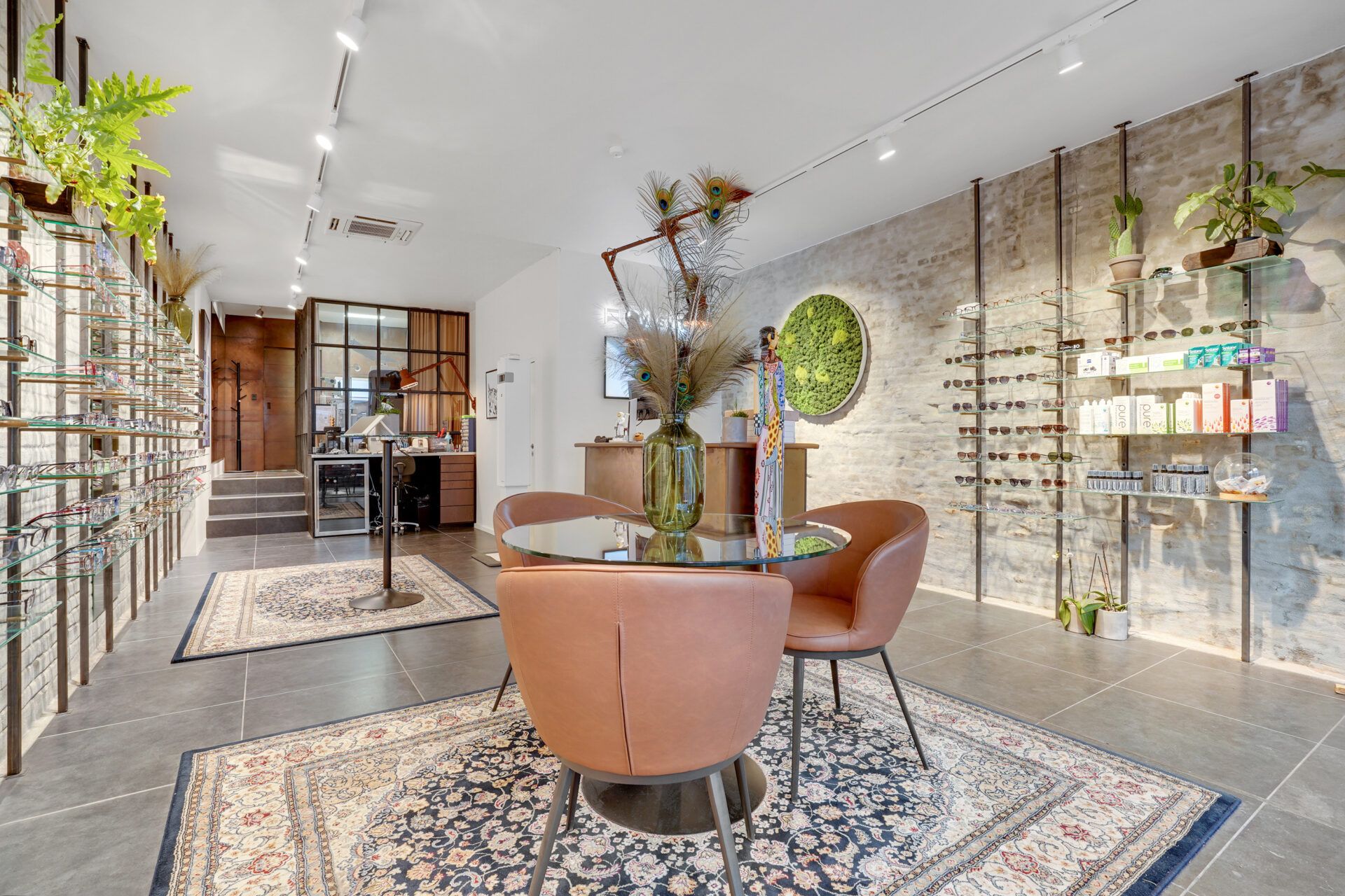 I en moderne butik med lyse gulve og vægge står et rundt glasbord med to brune stole og en grøn vase med fjer i midten.