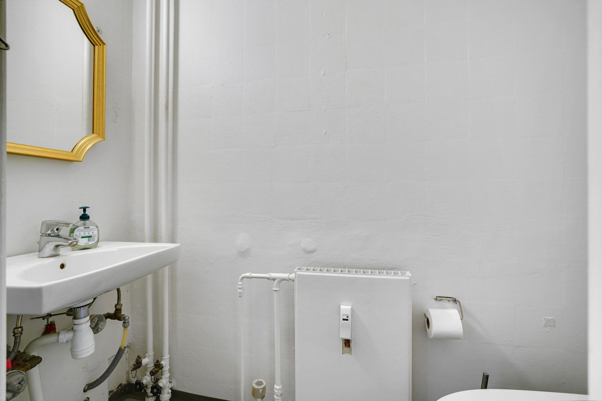 En lille hvid badeværelse med et badekar, håndvask og håndklædeholder samt et guldbelagt spejl på væggen. Der er også en