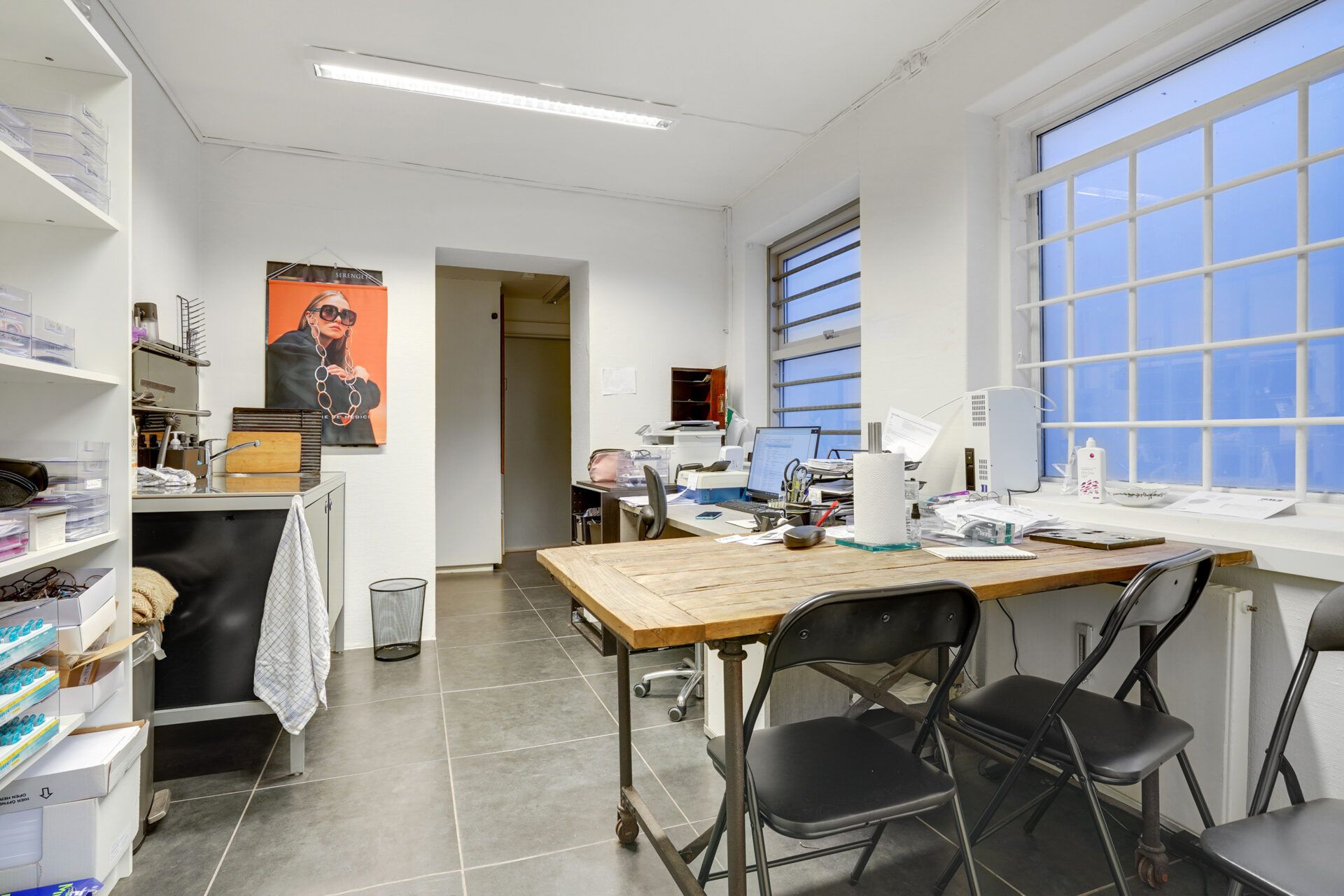 Foto af et lille kontorrum med tre stole, et bord, en computer, en faxmaskine og et skab. Væggene er hvide, og der er en høj