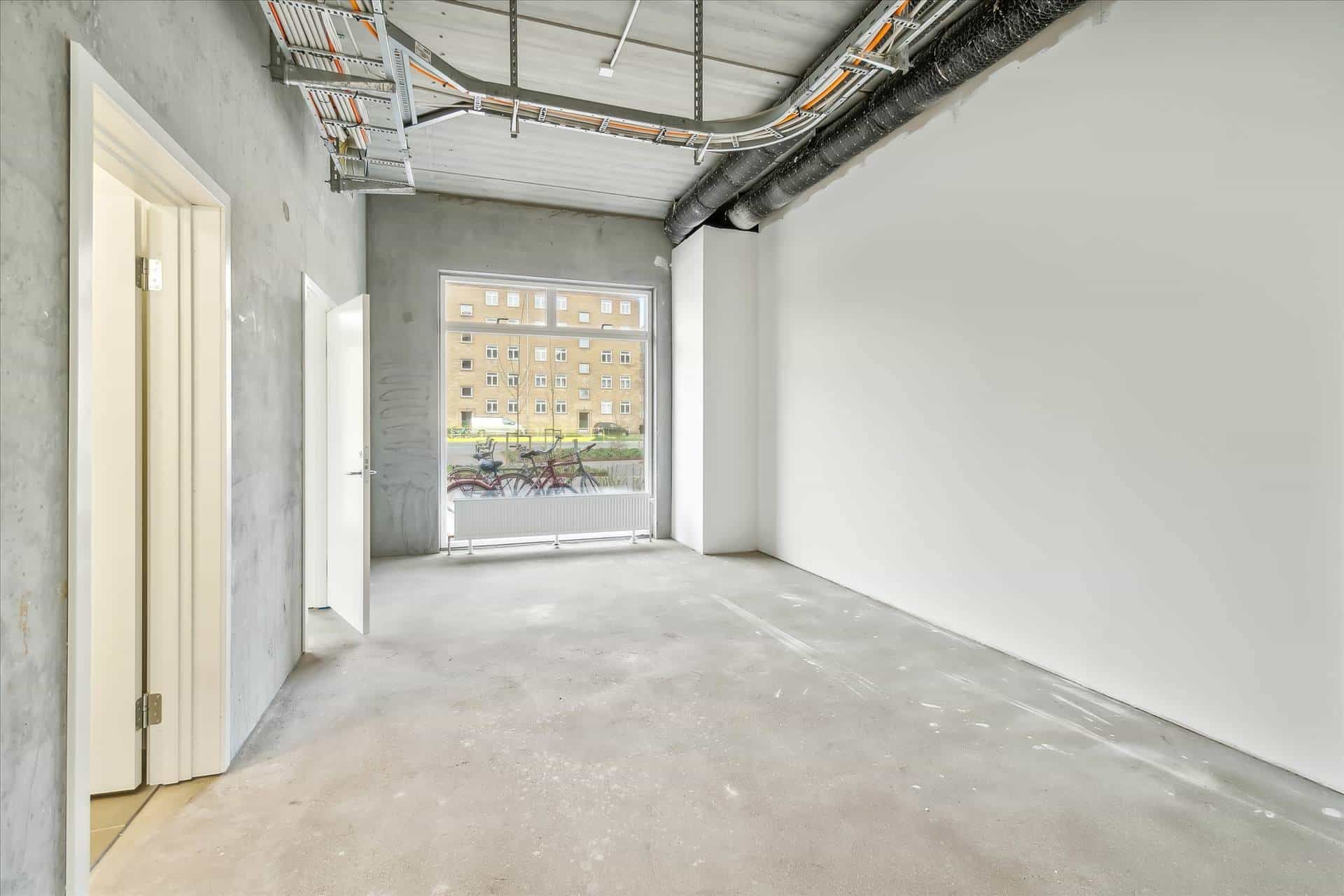 En stor, tom værkstedslokal med hvid gulv, hvide vægge og åbne ruder med udsigt til bygninger og cykler i baggrunden.