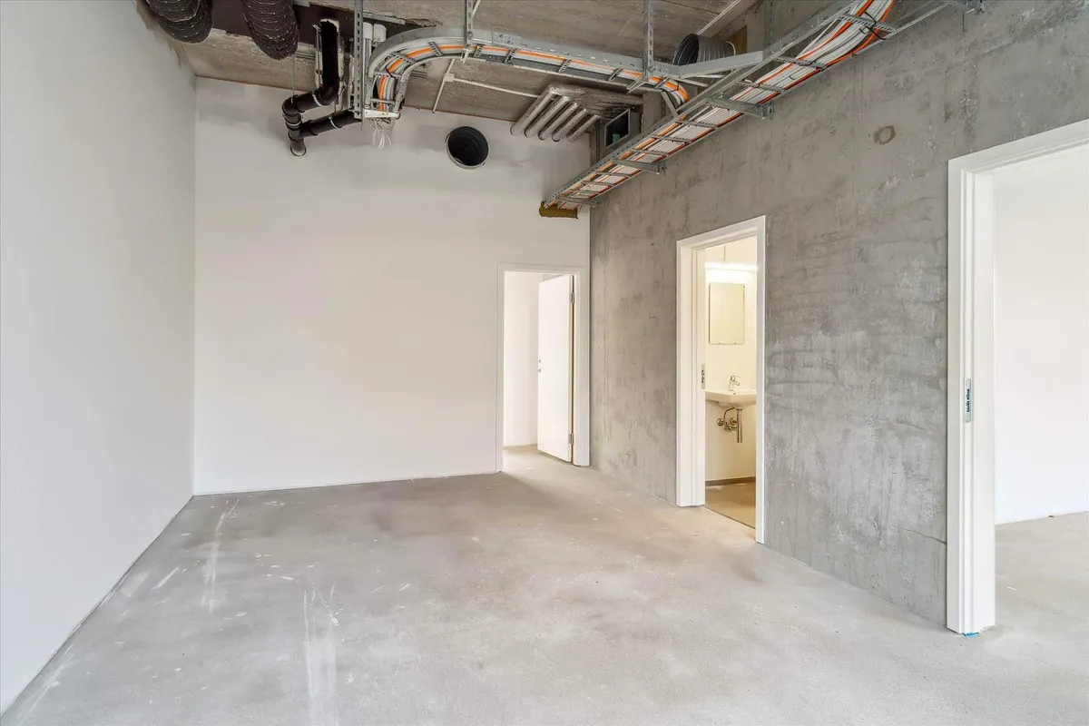 Et stort, tomt værelse med hvidt gulv og betonvægge samt flere åbninger til andre værelser og tekniske faciliteter som