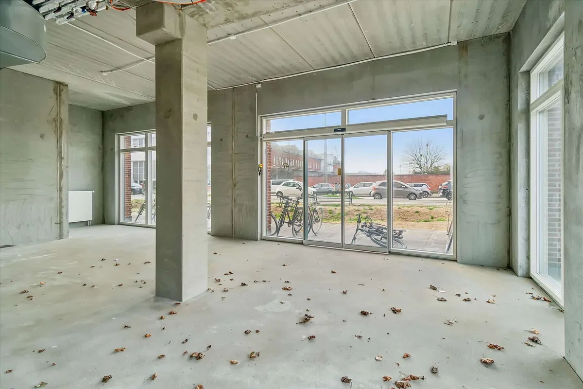 Tomt værelse med glasdøre og vinduer samt tre cykler stillet udenfor. Rummet er fuld af løv, og der er biler parkeret på