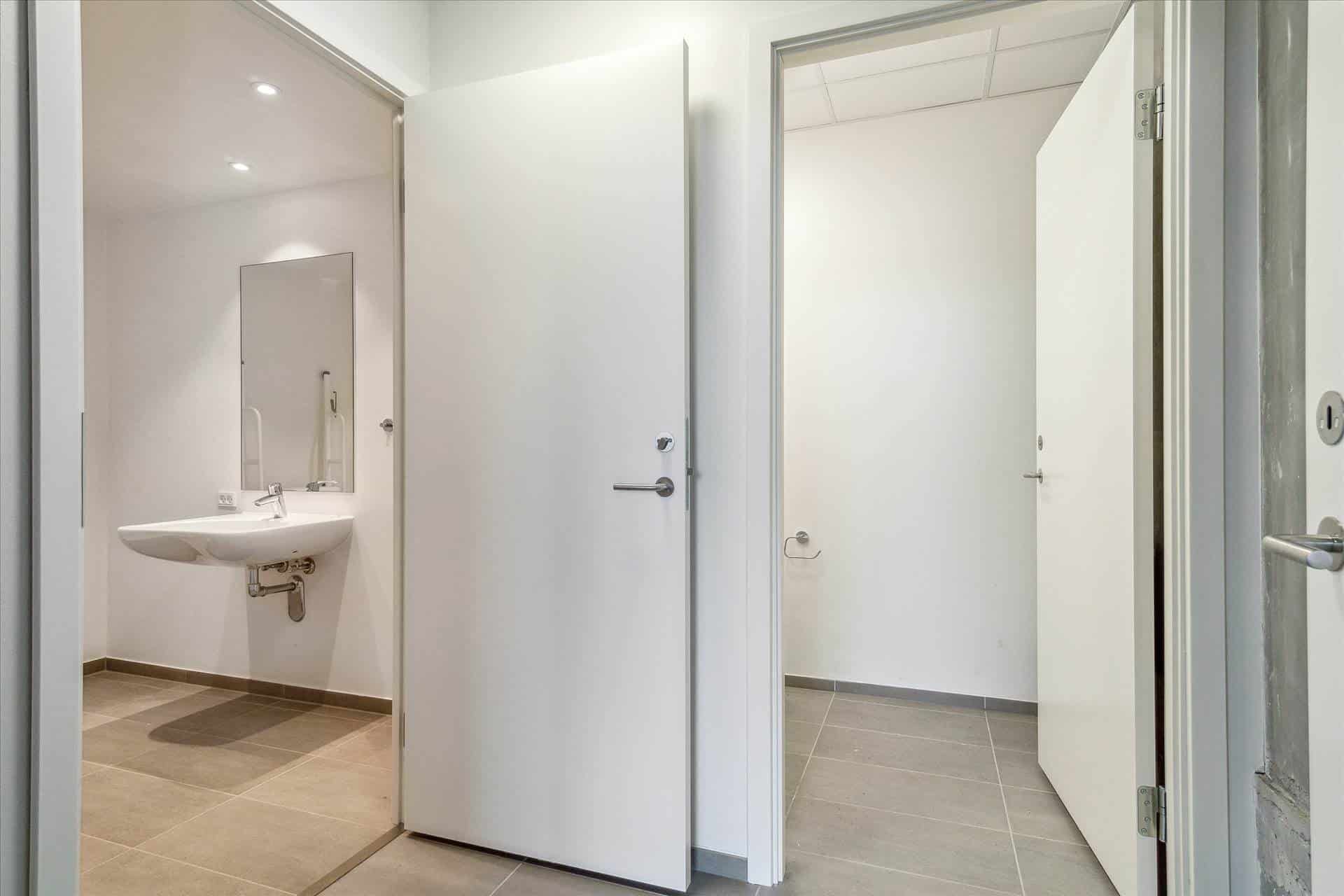 En moderne hvid badeværelsesdør er åben og viser et fladt toilet med en hvid vask og spejl samt et håndklædeholder på væggen.