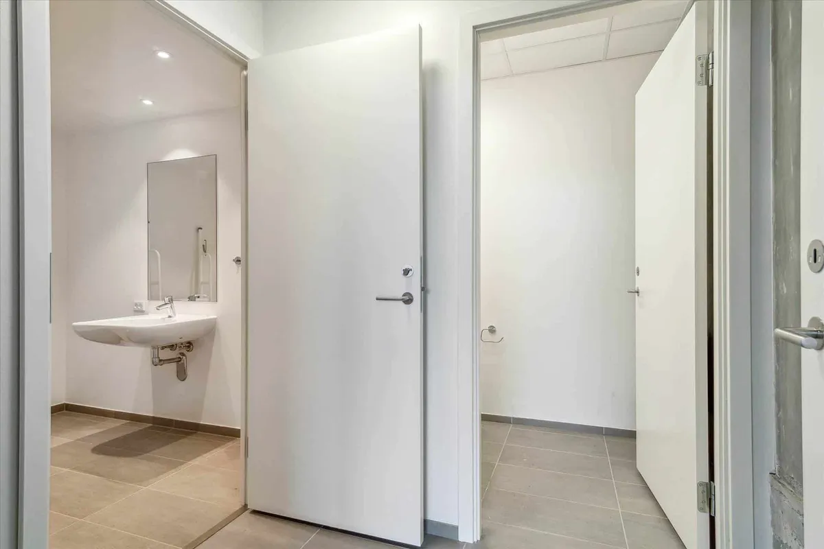 En moderne hvid badeværelsesdør er åben og viser et fladt toilet med en hvid vask og spejl samt et håndklædeholder på væggen.