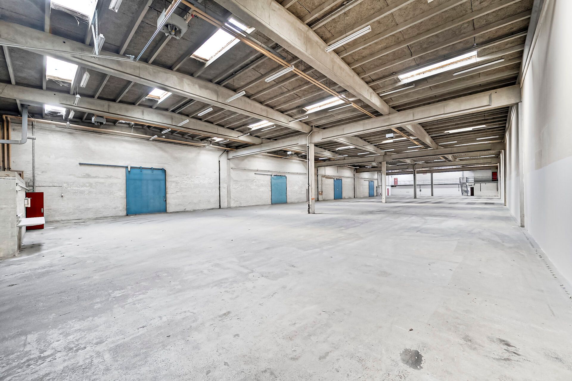 En stor lagerhal med cementgulve og lyser. Der er flere blå porte og røde døre, samt flere lysekroner.