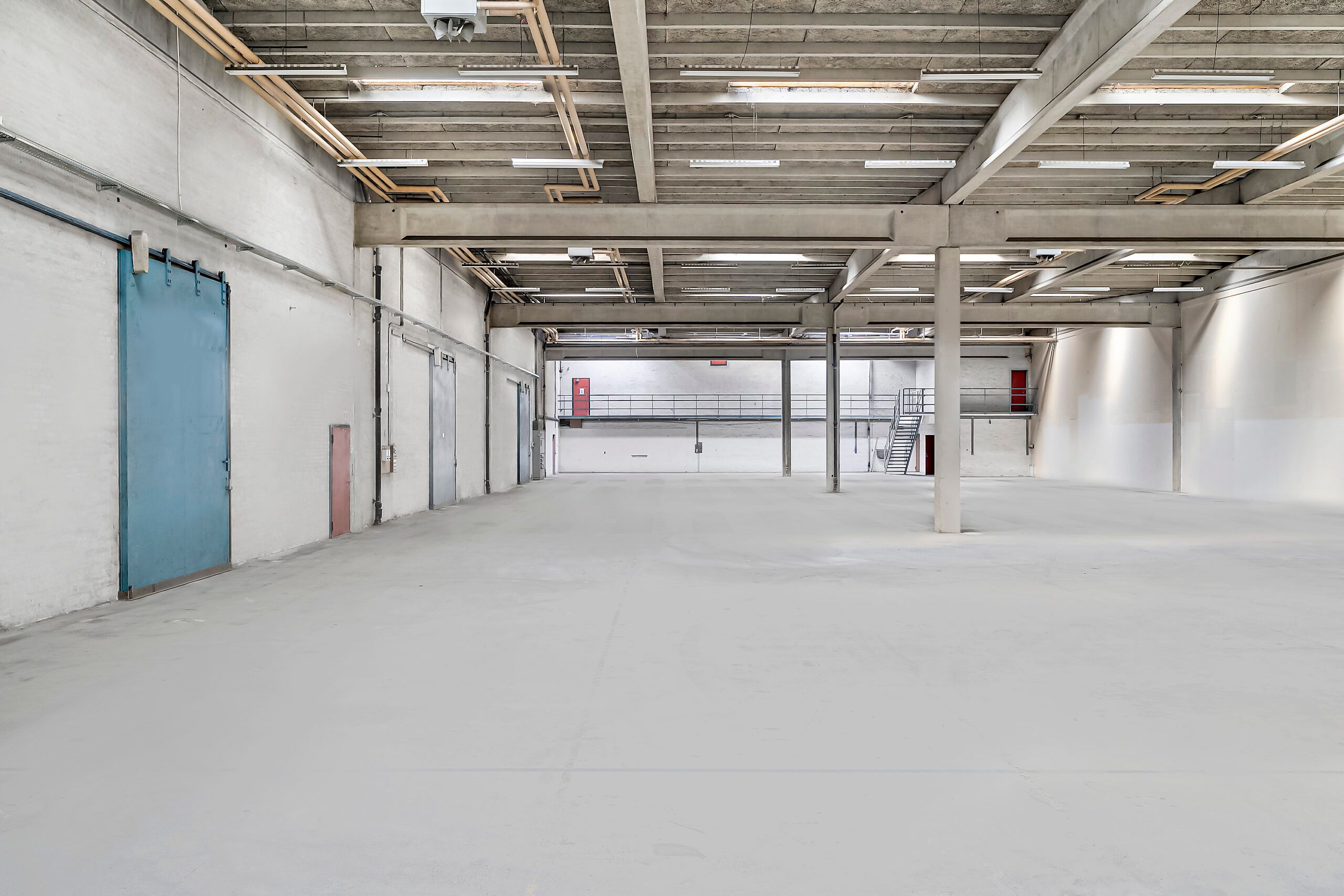 Lager med loftsbjælker og gulv med lyse armaturer, høje vinduer, røde døre og lyseblå sektionsdør til venstre.