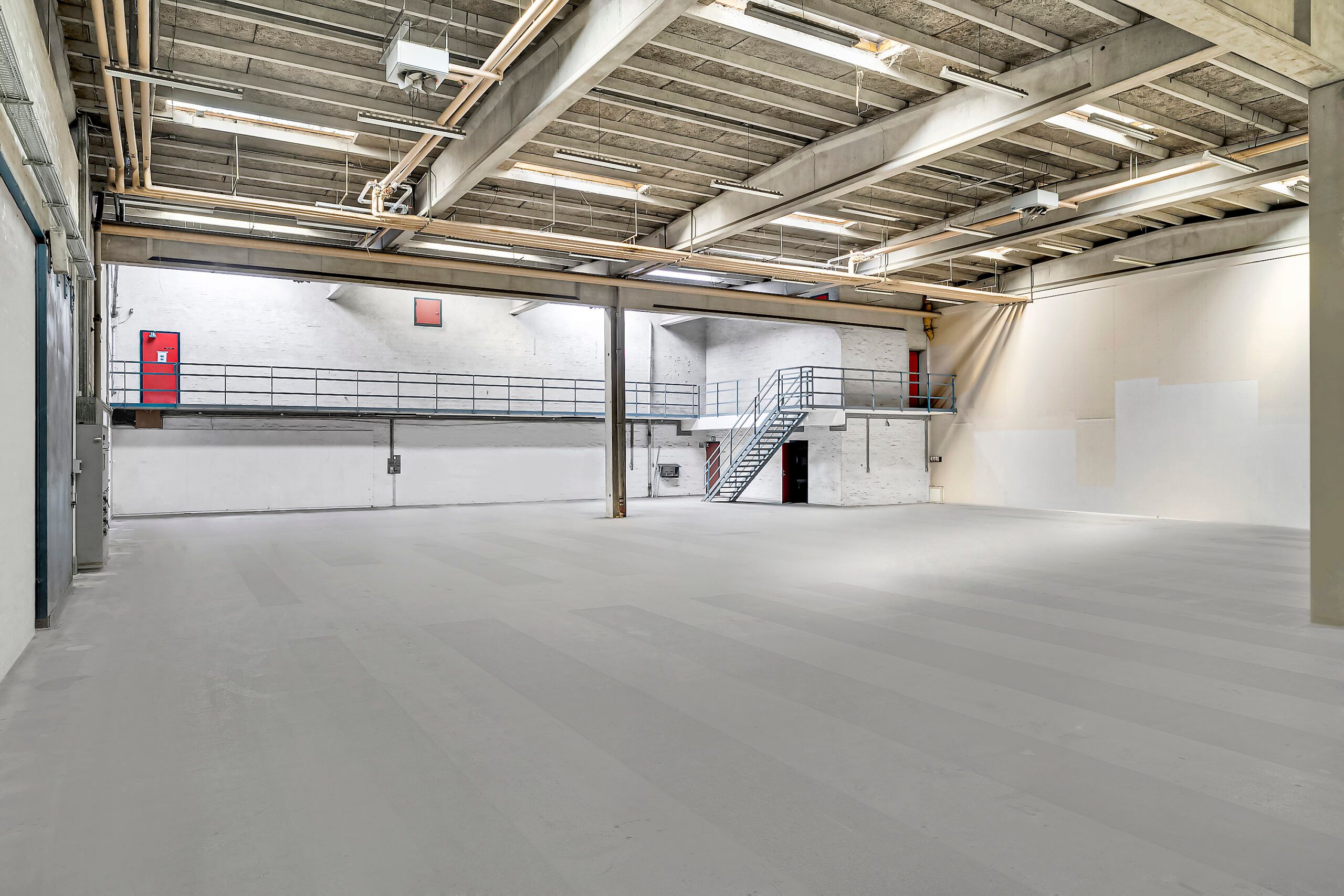 En stor industrihal med højt loft, metalrammer og to gangbroer med trapper, der fører til en mellemgulv. Der er en rødt dør