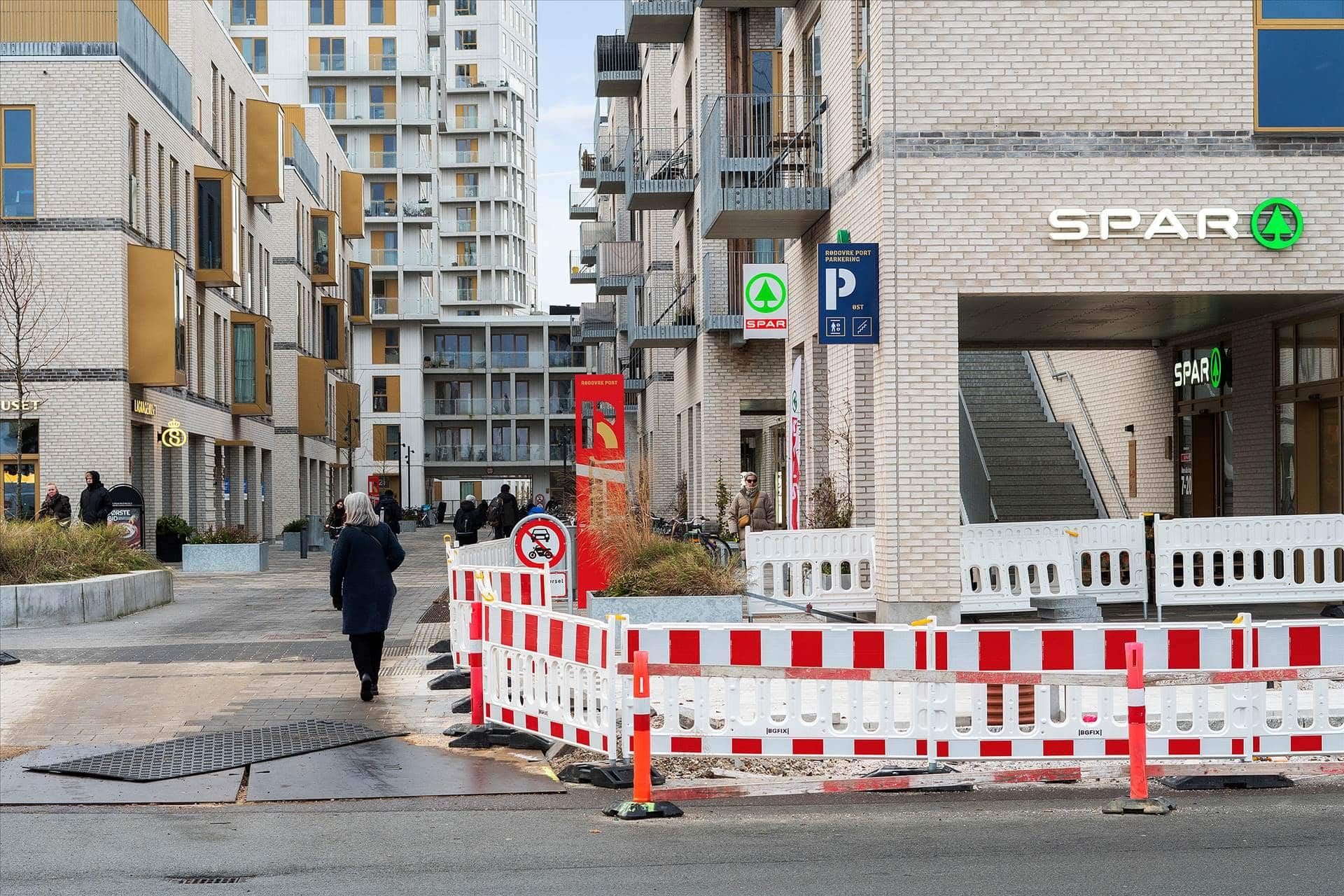 En gågade med forhøjede områder, røde og hvide barrierer og SPAR-skilte ved en bygning med adgangstrapper og et stort rødt