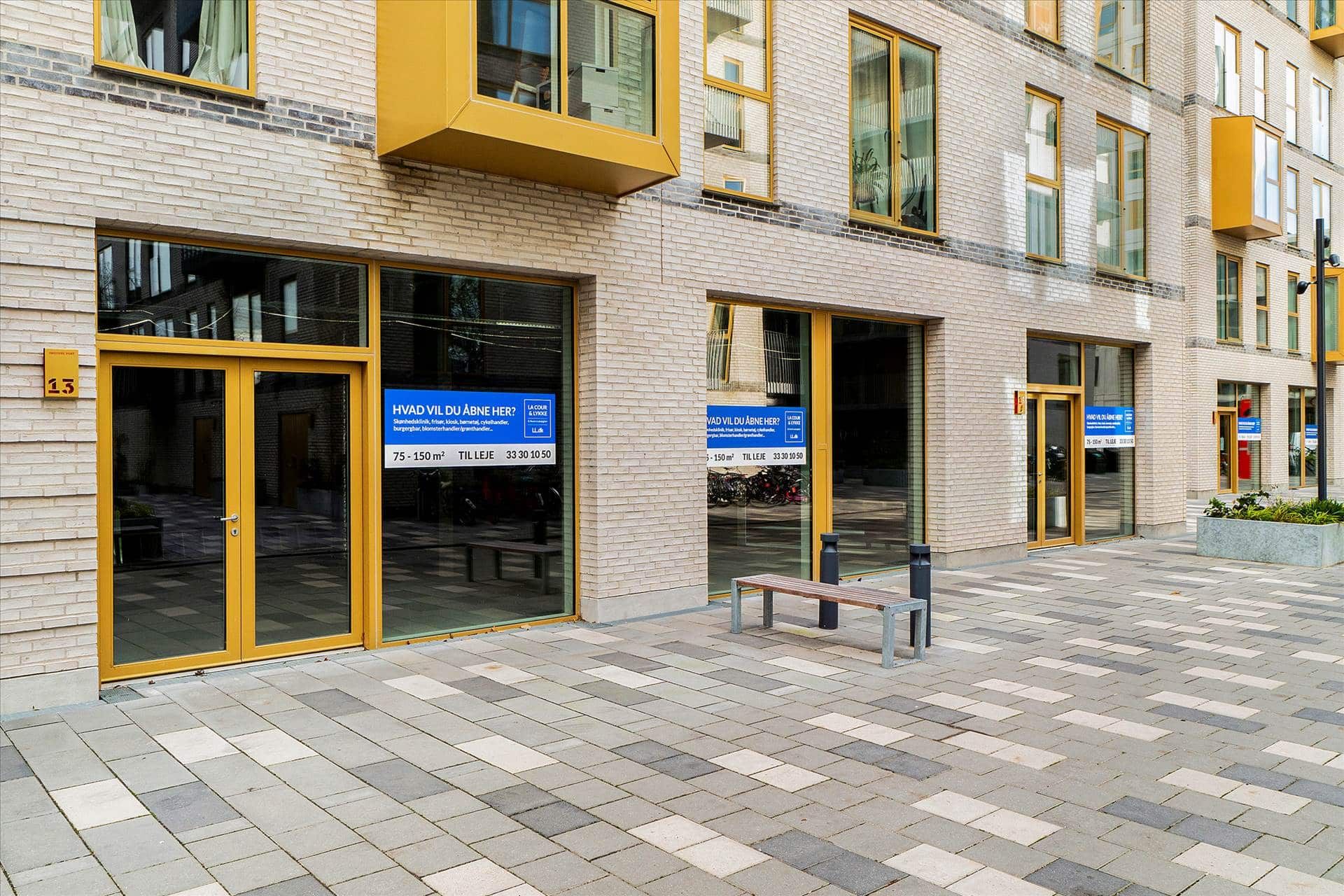 Butikslokaler med gule døre og vinduer står på hjørnet af en gade med en fortov. Foran dørene er der et benk og annoncer med