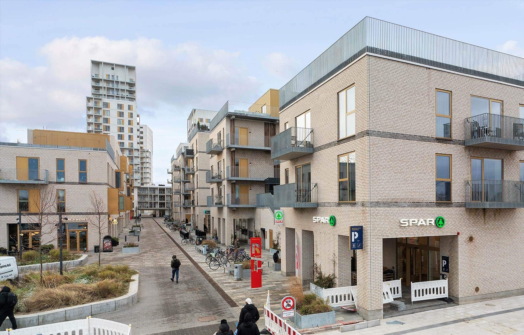 Mennesker går langs gågaden i Ørestad med spar butik og parkeringsplads og højhus bagved. Foran er buske og et hvidt