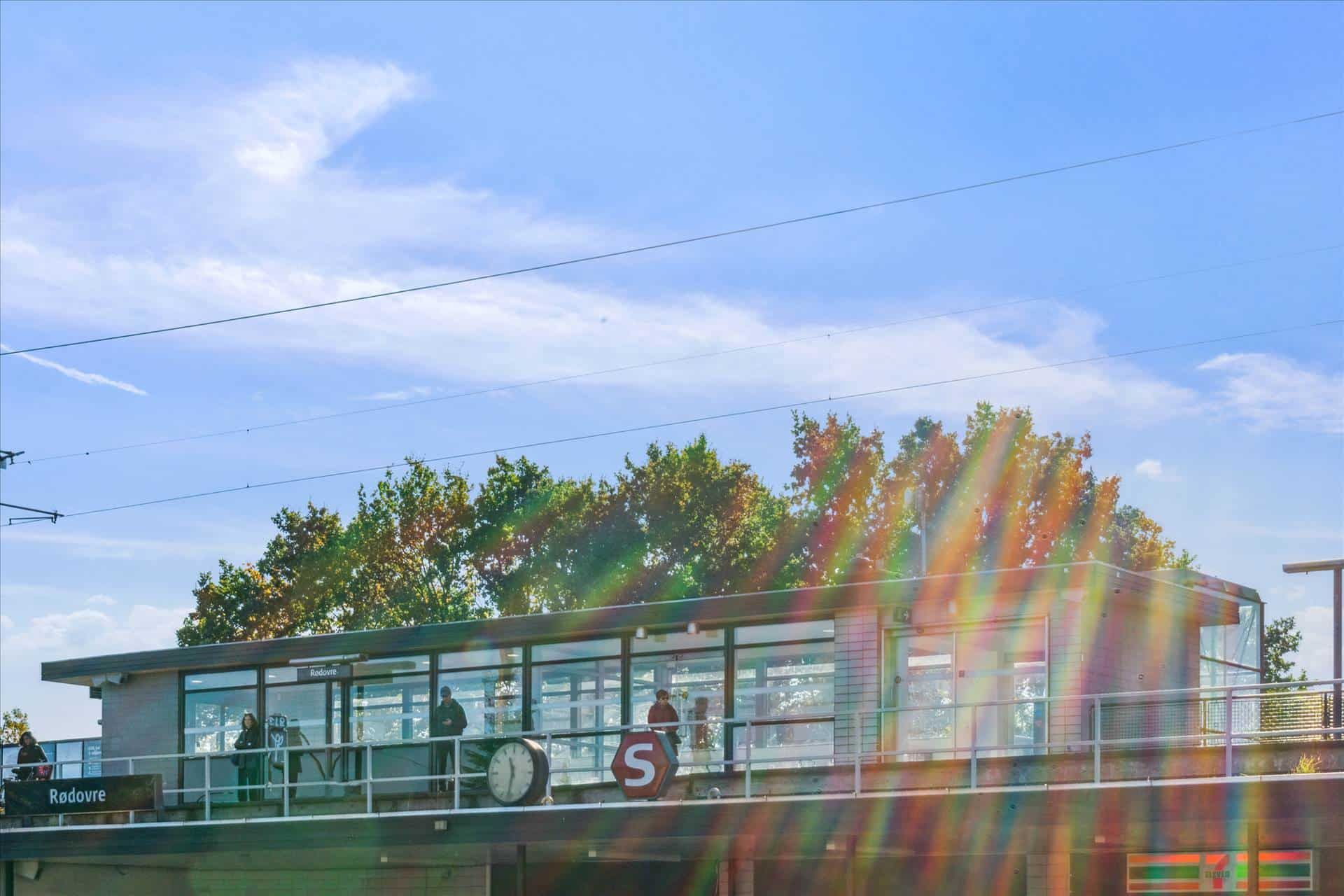 Folk står på en platform med en s-skilt, et ur og en udsigt over Rodovre og en del af skoven i baggrunden