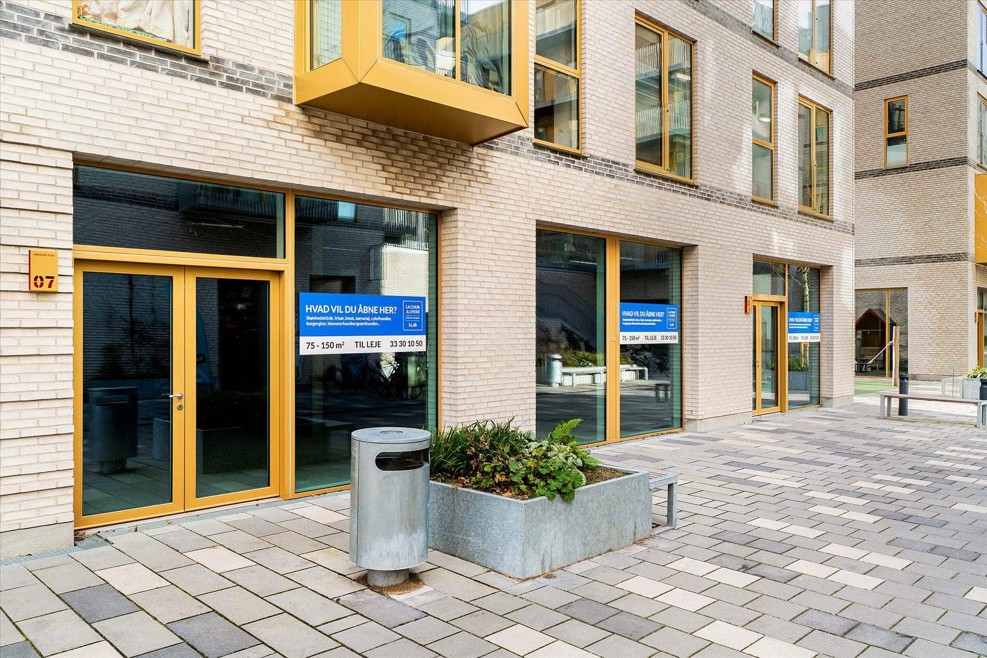 Ledig lokale i bygningen med indskrift på vinduet. Der er skraldespand og plantebed ved siden af. En stol og høj bygning i