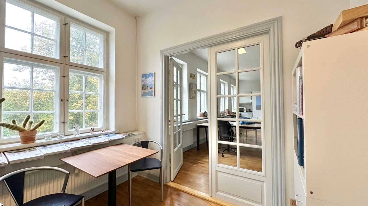 Inde i et studierum med trægulv og højt loft står et bord og to stole med udsigt til haven